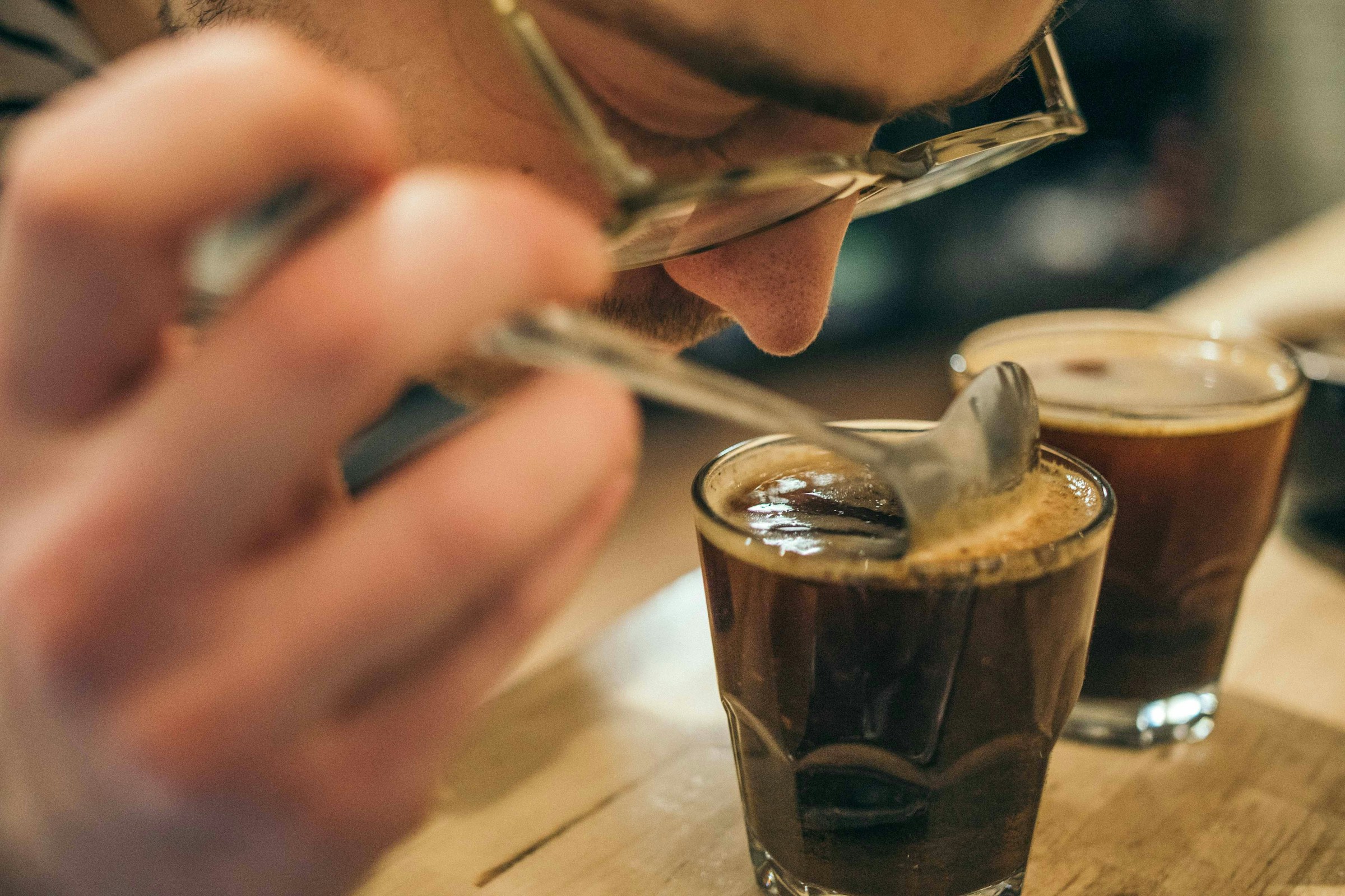 A person wearing glasses leans close to a glass of espresso as if smelling it.