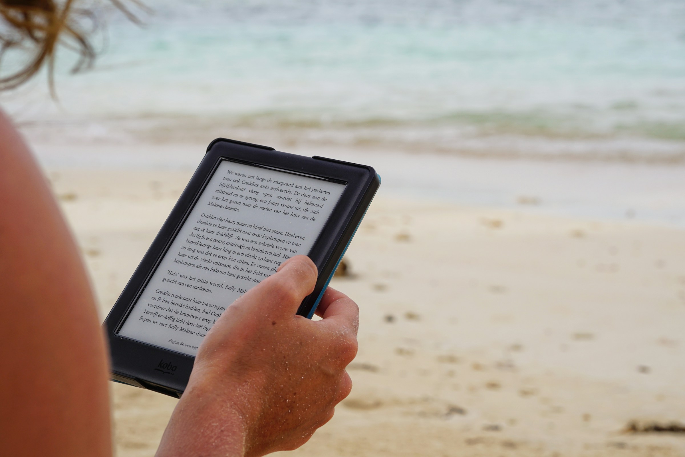 Over-the-shoulder view of a person sitting on the beach, reading a story on an e-reader.