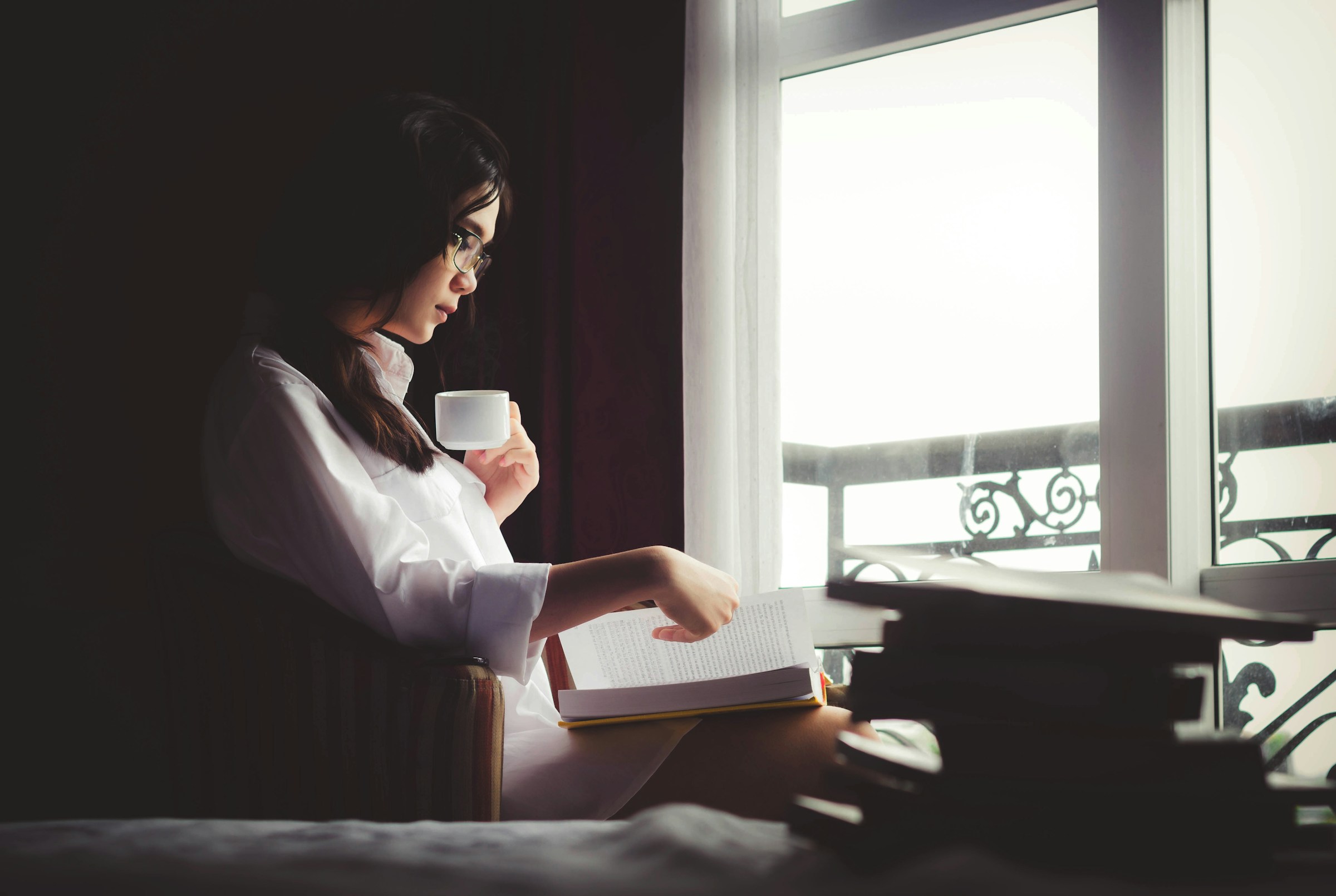 A person sits in front of a window in a dark room, drinking from a white mug and reading a book.