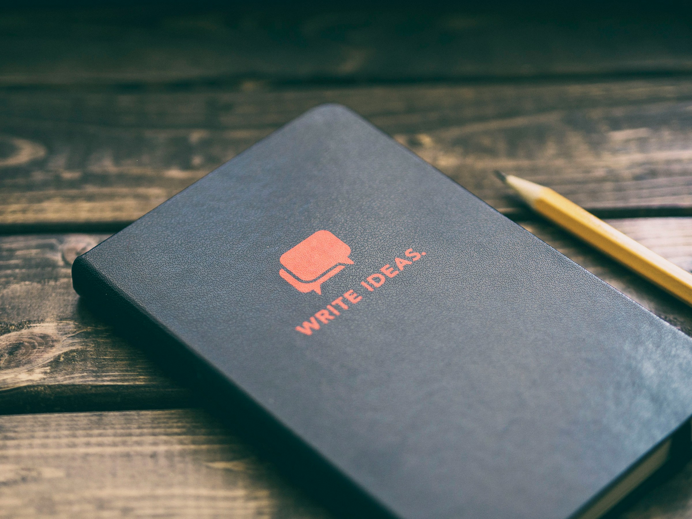 A black journal with "WRITE IDEAS" on the cover sits on a wooden surface beside a pencil.