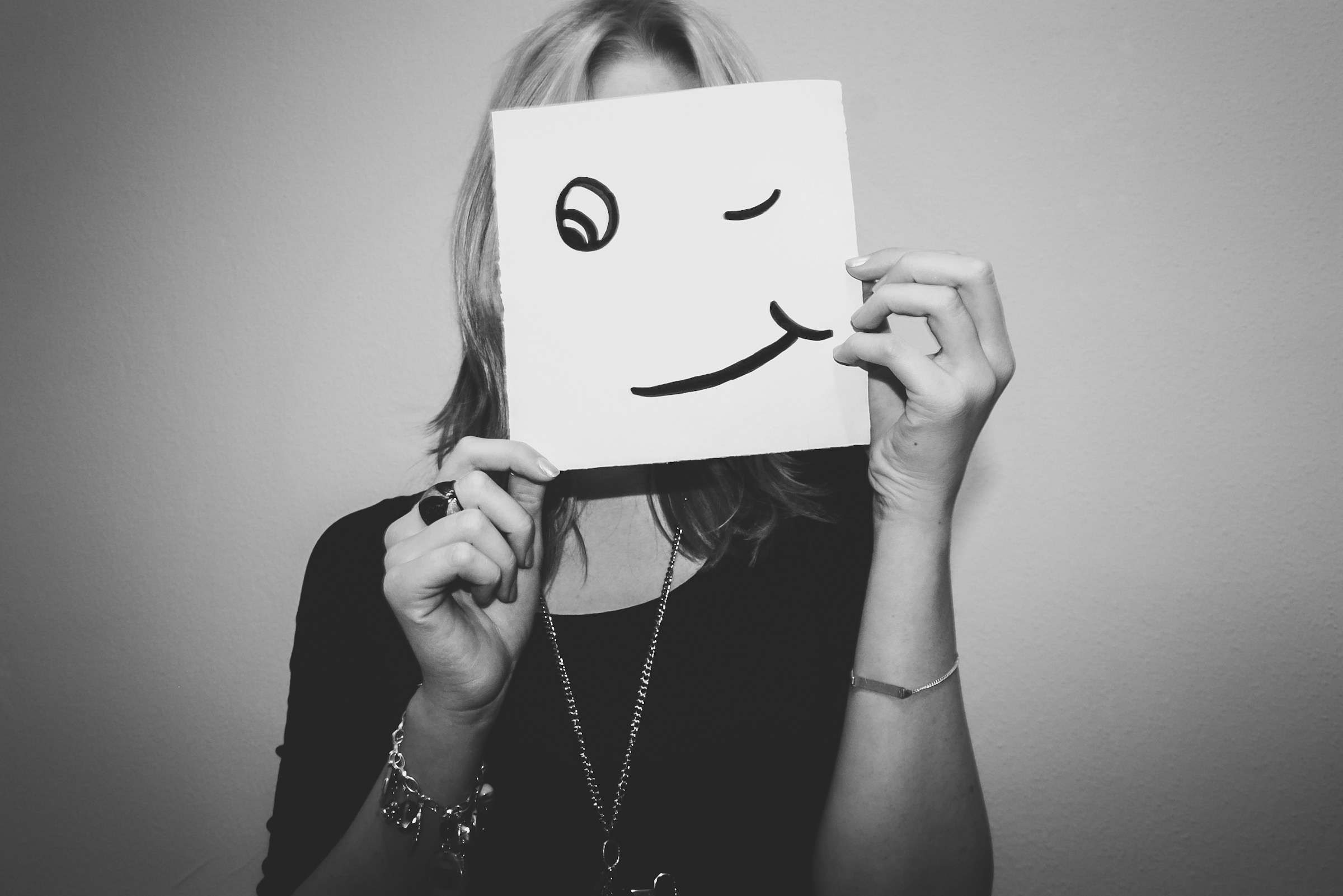 A person holds a paper with a winky face drawn on it in front of their actual face.