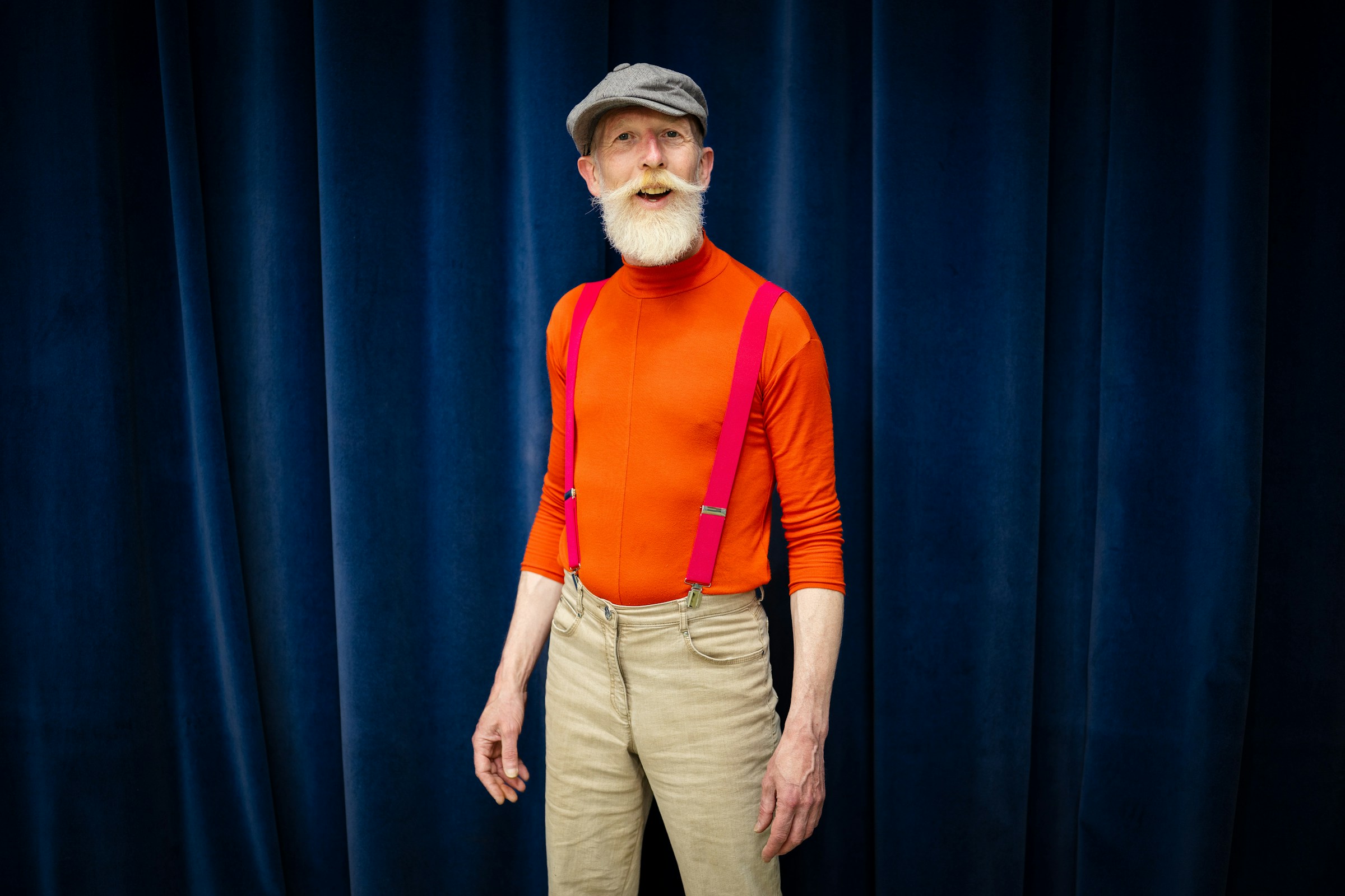 A person with a white beard, red shirt, red suspenders, and cap stands in front of a navy blue curtain.
