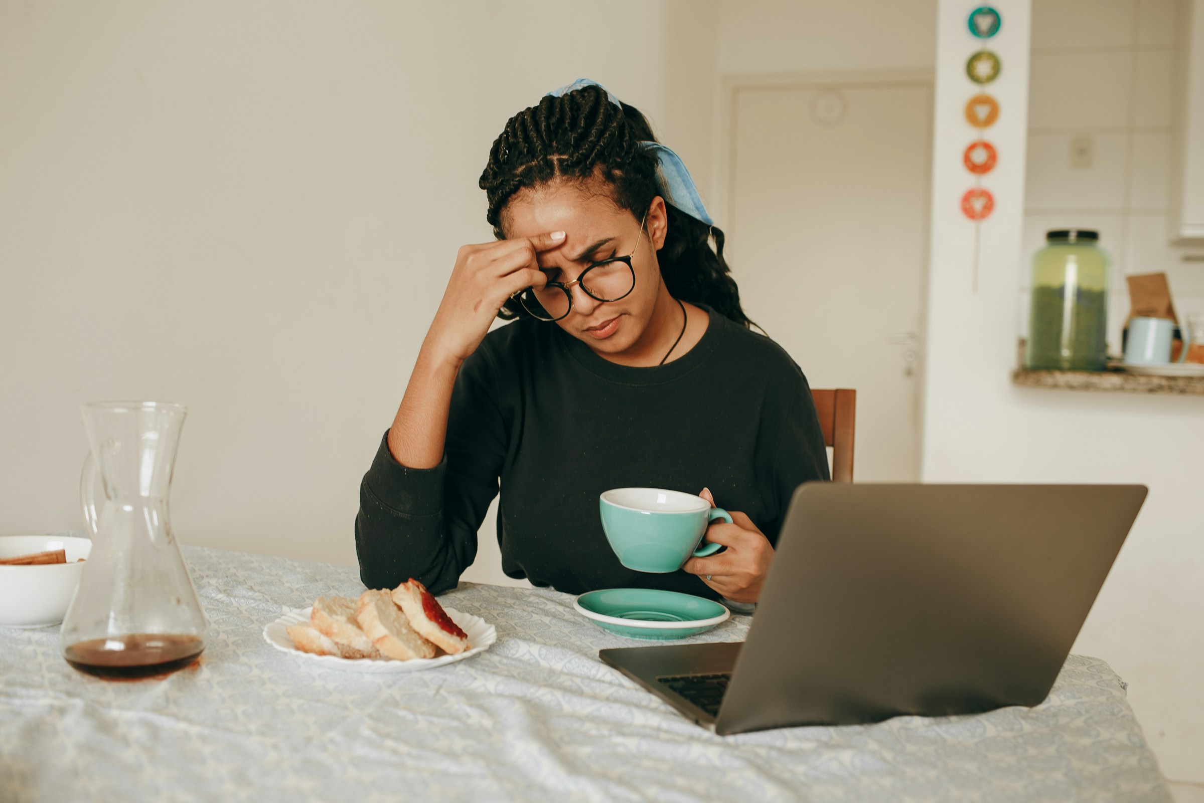 A person with glasses and a scarf in their hair sits at a breakfast table in front of an open laptop, holding a blue teacup and rubbing their forehead.