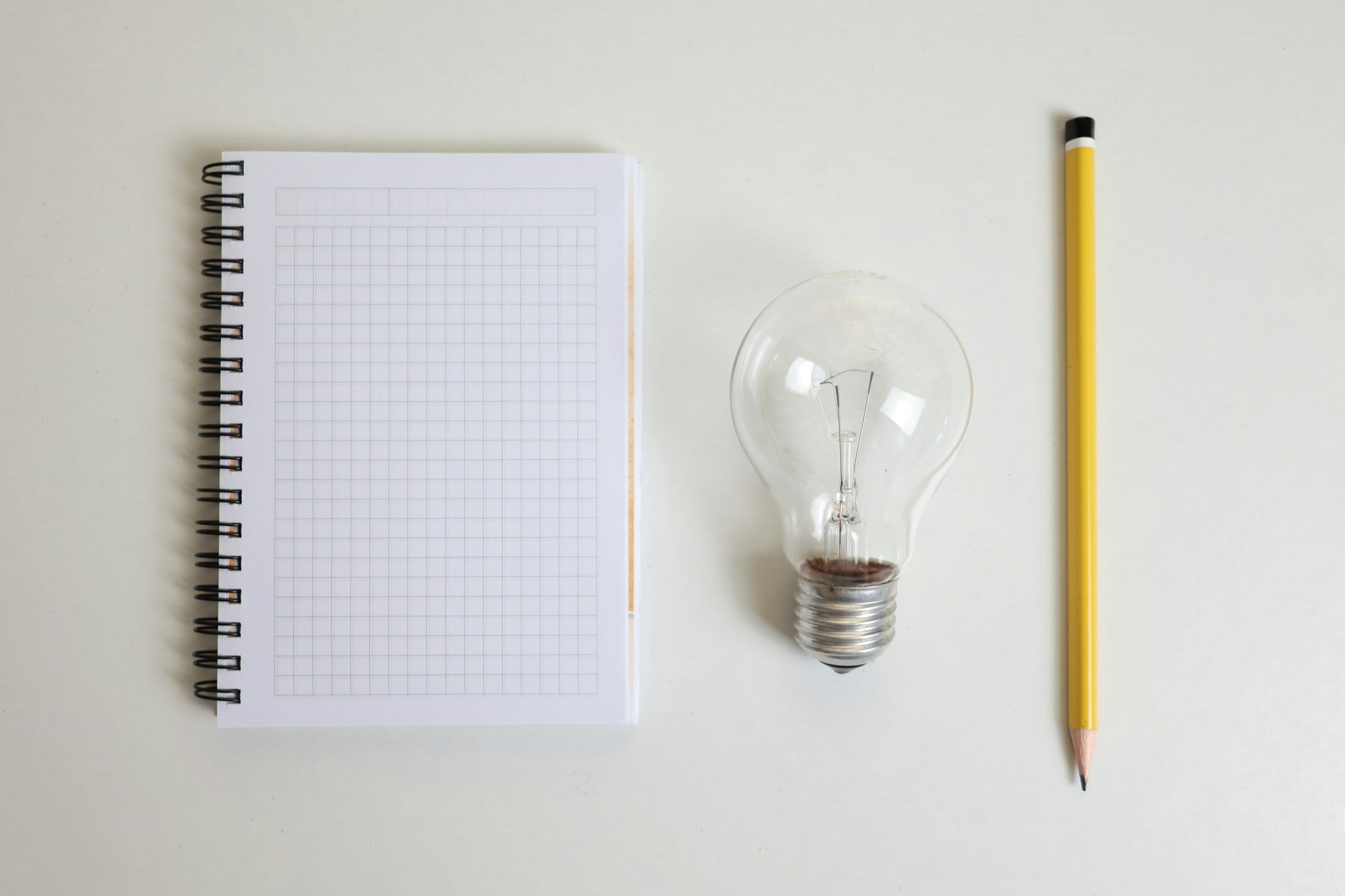 A blank notebook, a lightbulb, and a pencil sit in a row on a white surface.