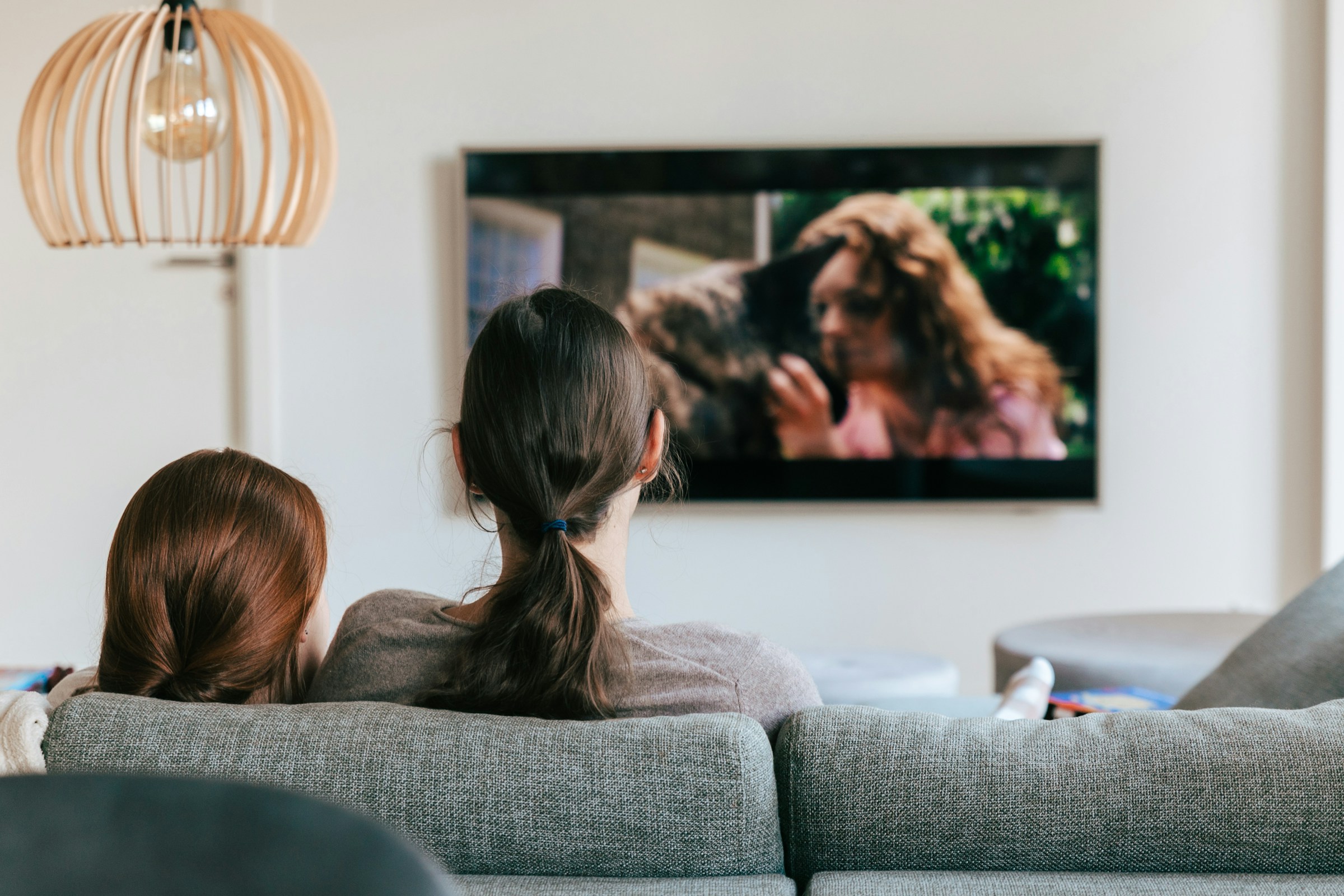 Two people with ponytails sit close together on a couch, watching a TV screen showing a woman and a dog.