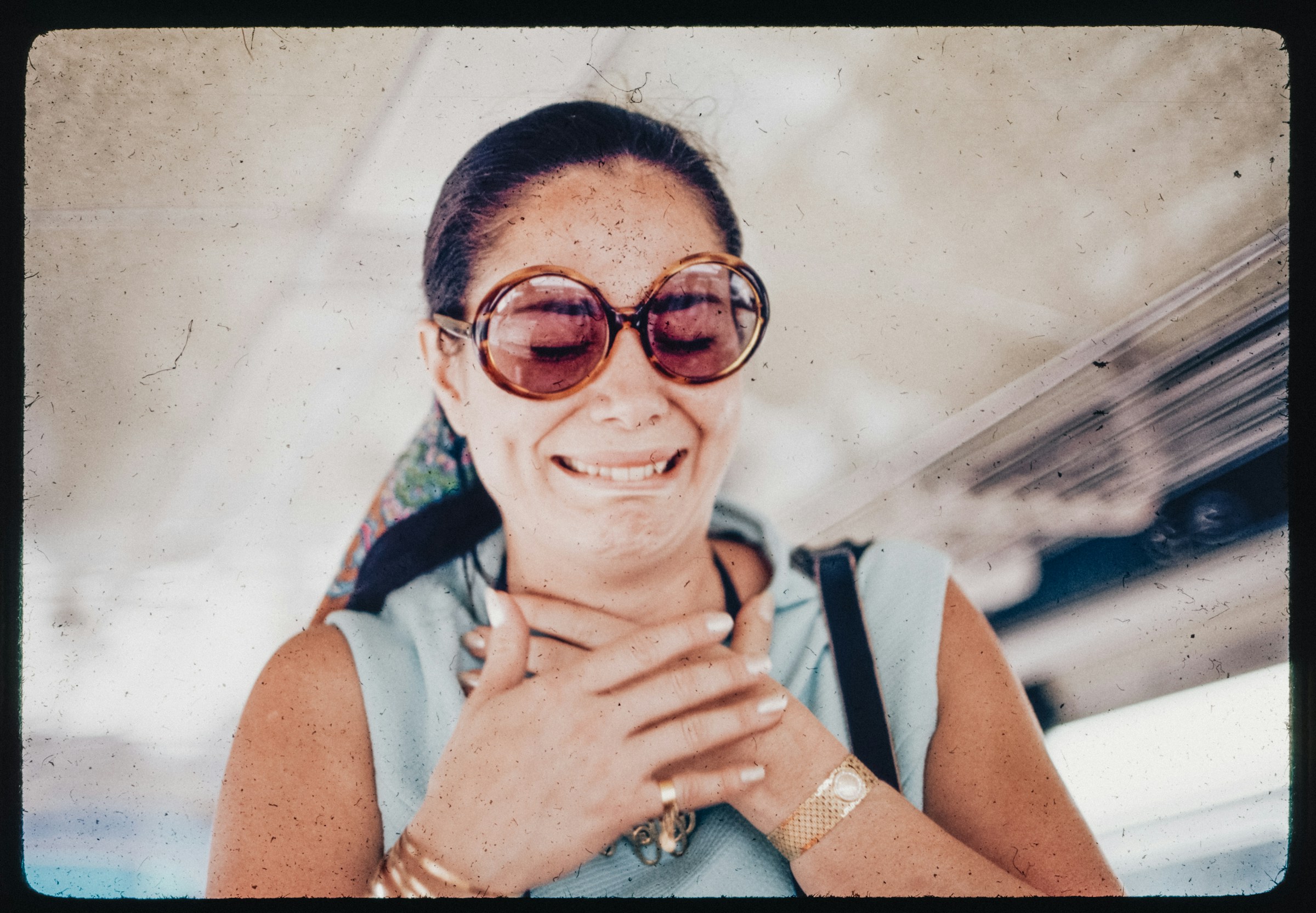 A person with a ponytail and big round tortoise shell sunglasses closes their eyes and grimaces while clutching their chest.