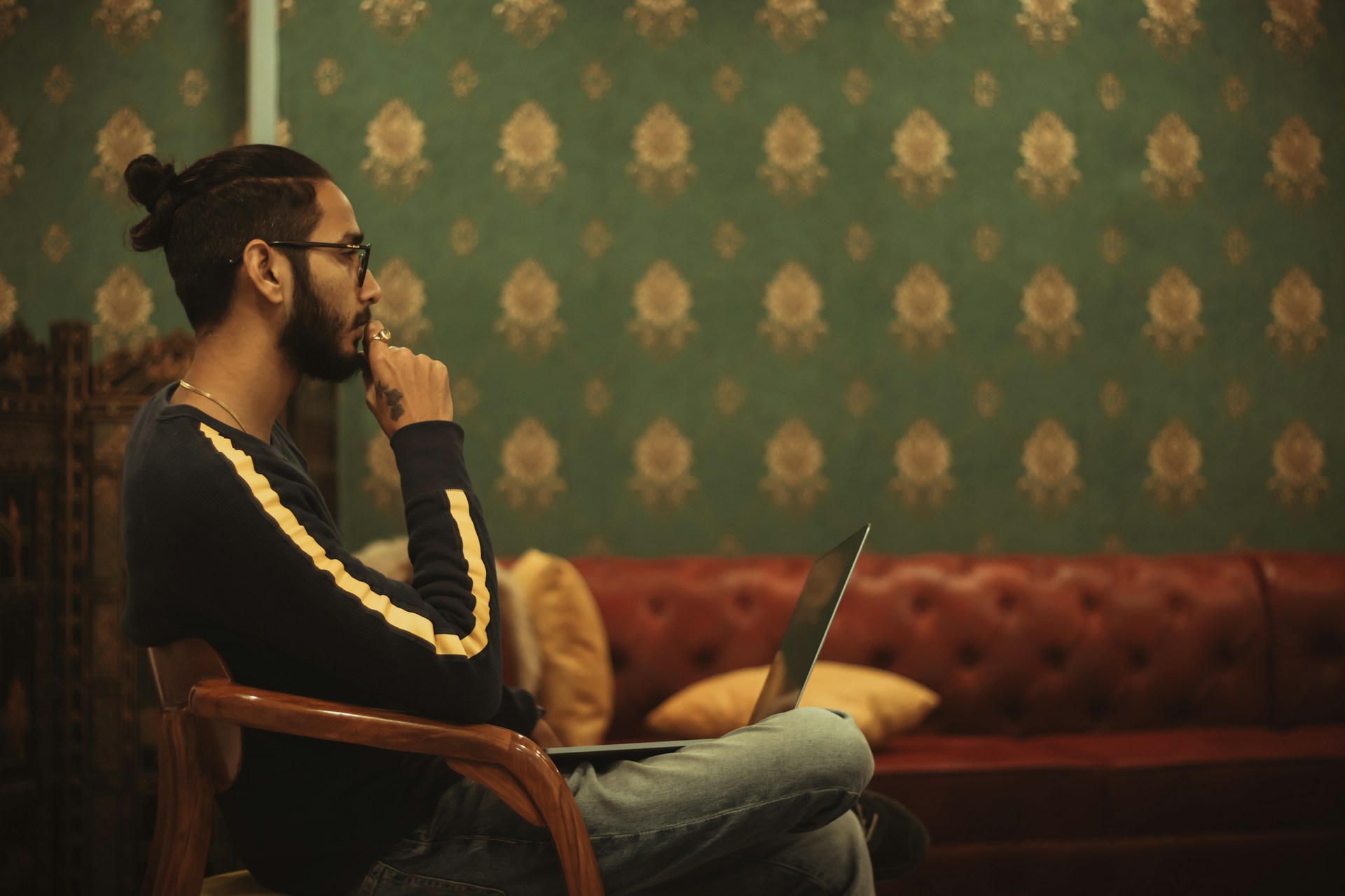 A person sits in a chair in a room with green and gold wallpaper. They hold a laptop in their lap and touch their finger to their mouth as they stare off into the distance.