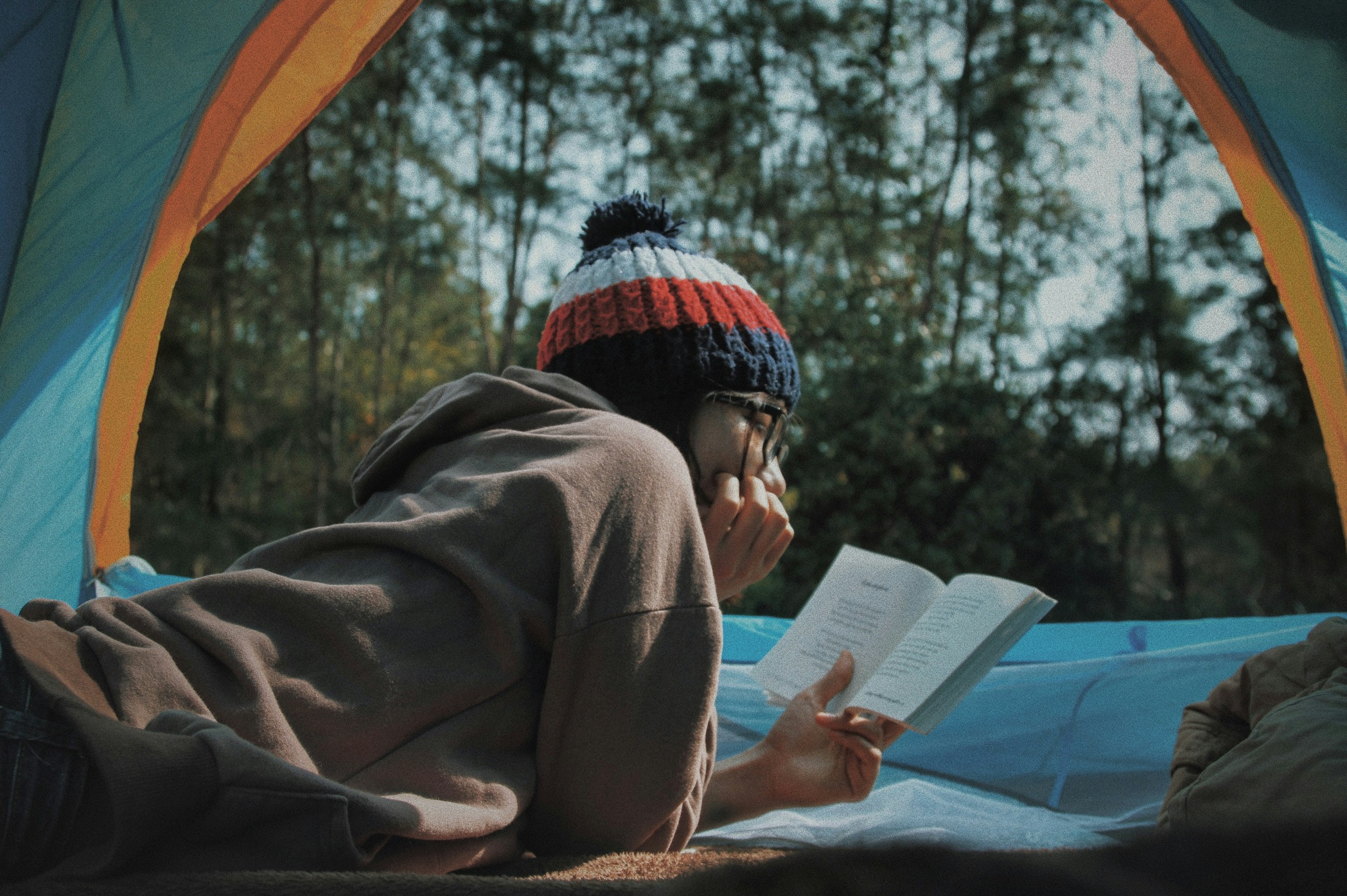 A person in glasses and a beanie lies on their stomach in a tent reading a book.