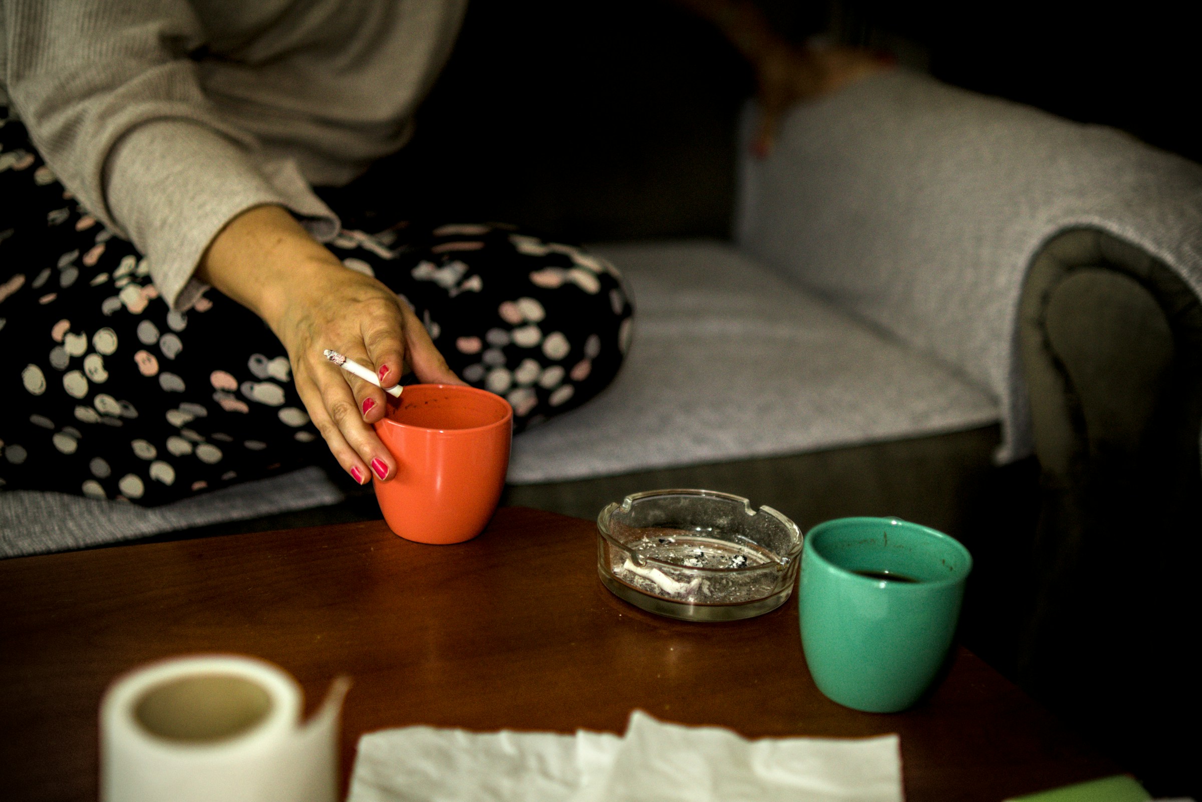A hand holding a cigarette reaches for a cup of coffee.
