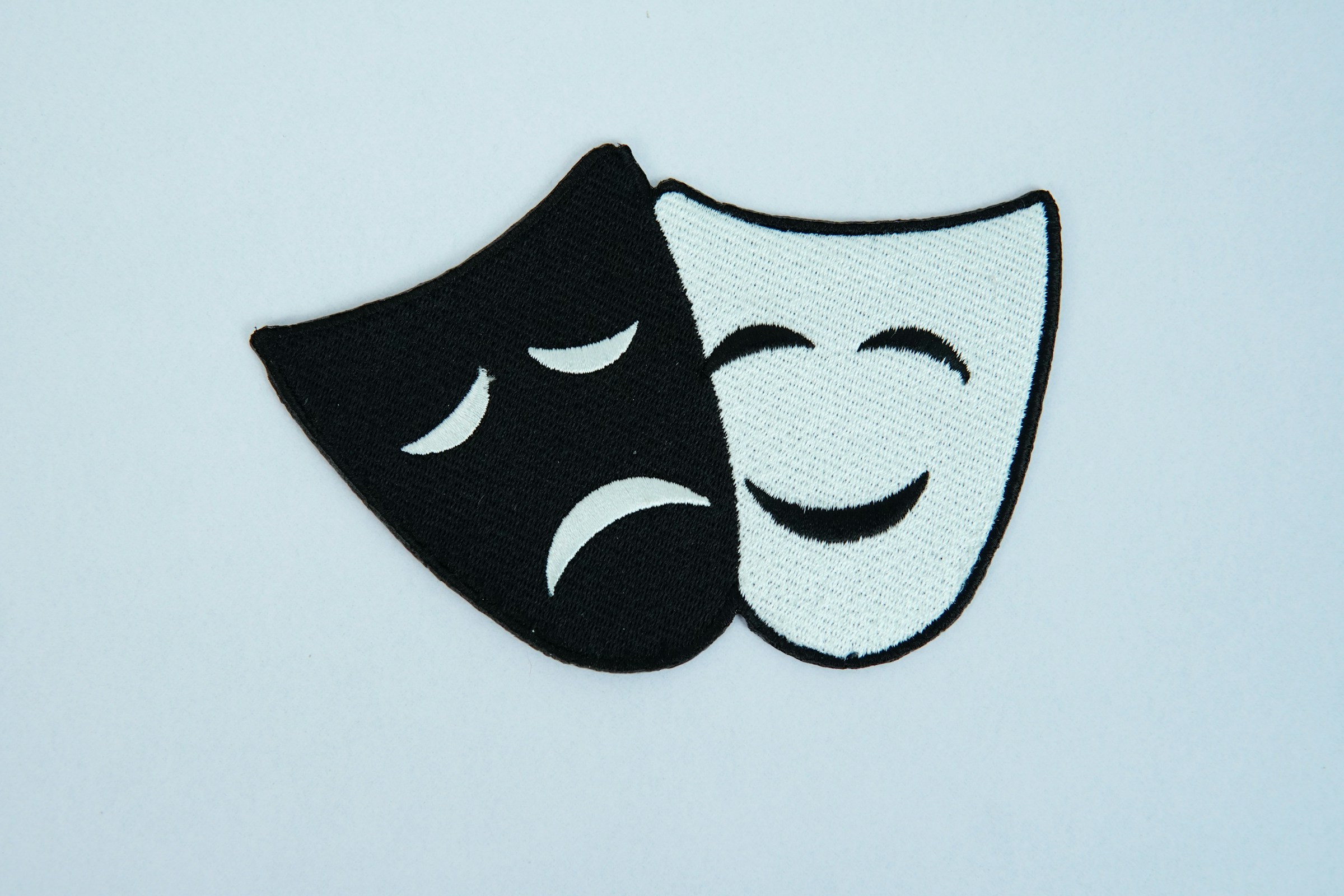 An embroidered black and white image of drama masks, one tragic and one comedic.