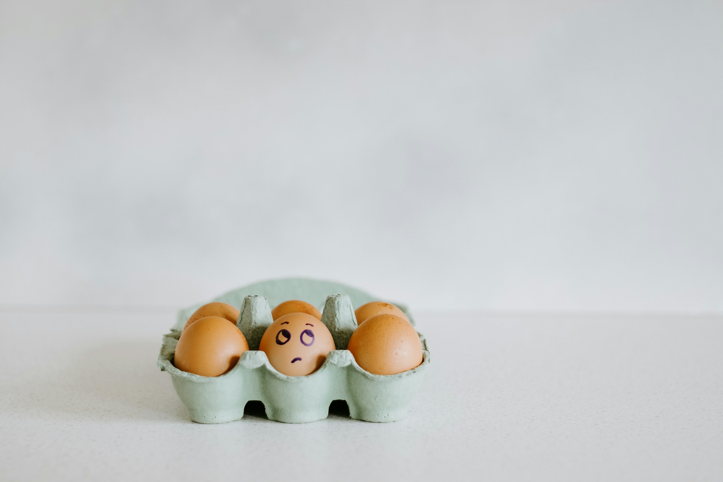Six brown eggs in a carton. One has an eyeroll face drawn on it.