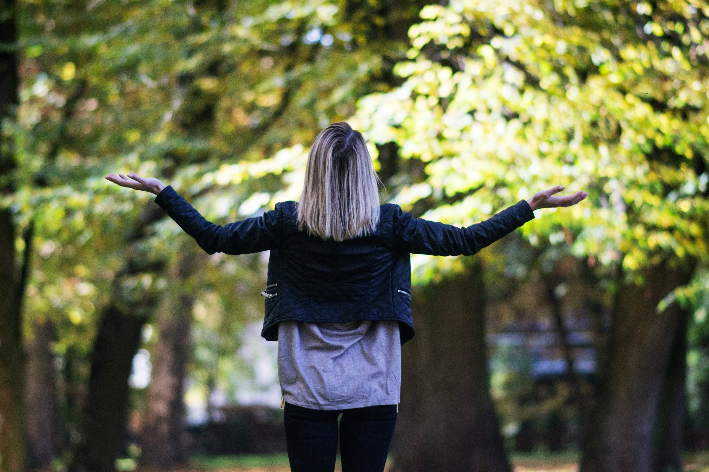 A person with shoulder-length blonde hair stands with their back to the camera and holds their palms up to the sky.