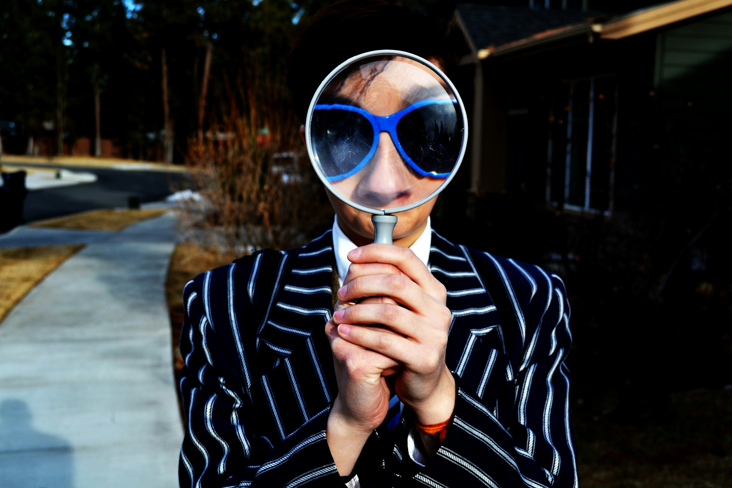 A person in a pinstriped blazer and blue sunglasses holds a magnifying glass up to their face.