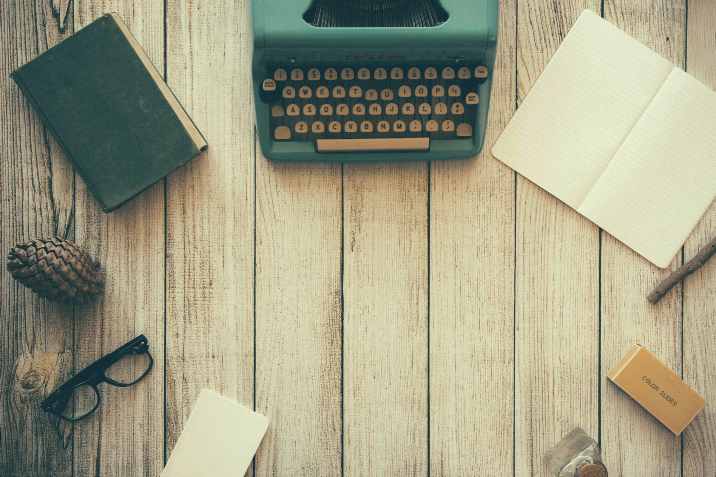 A green vintage typewriter, open notebook, closed book, pinecone, and glasses all arranged in a circle on a wood-paneled surface.