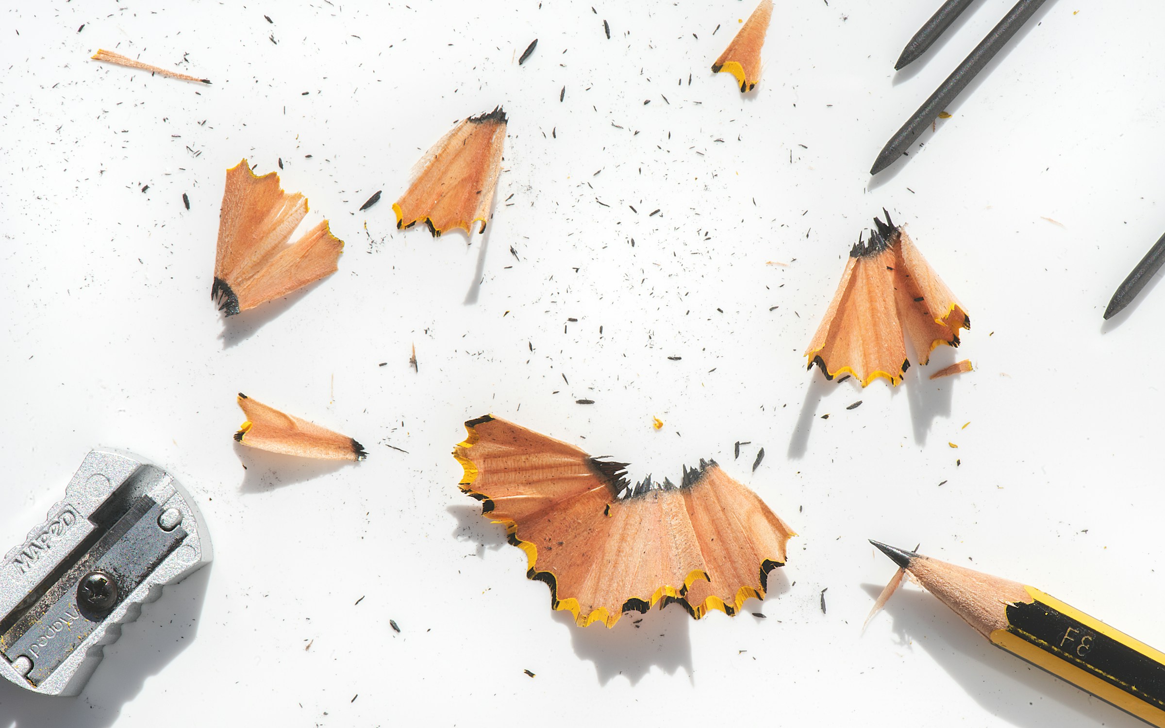 A sharp pencil, a pencil sharpener, and pencil shavings on a white surface.