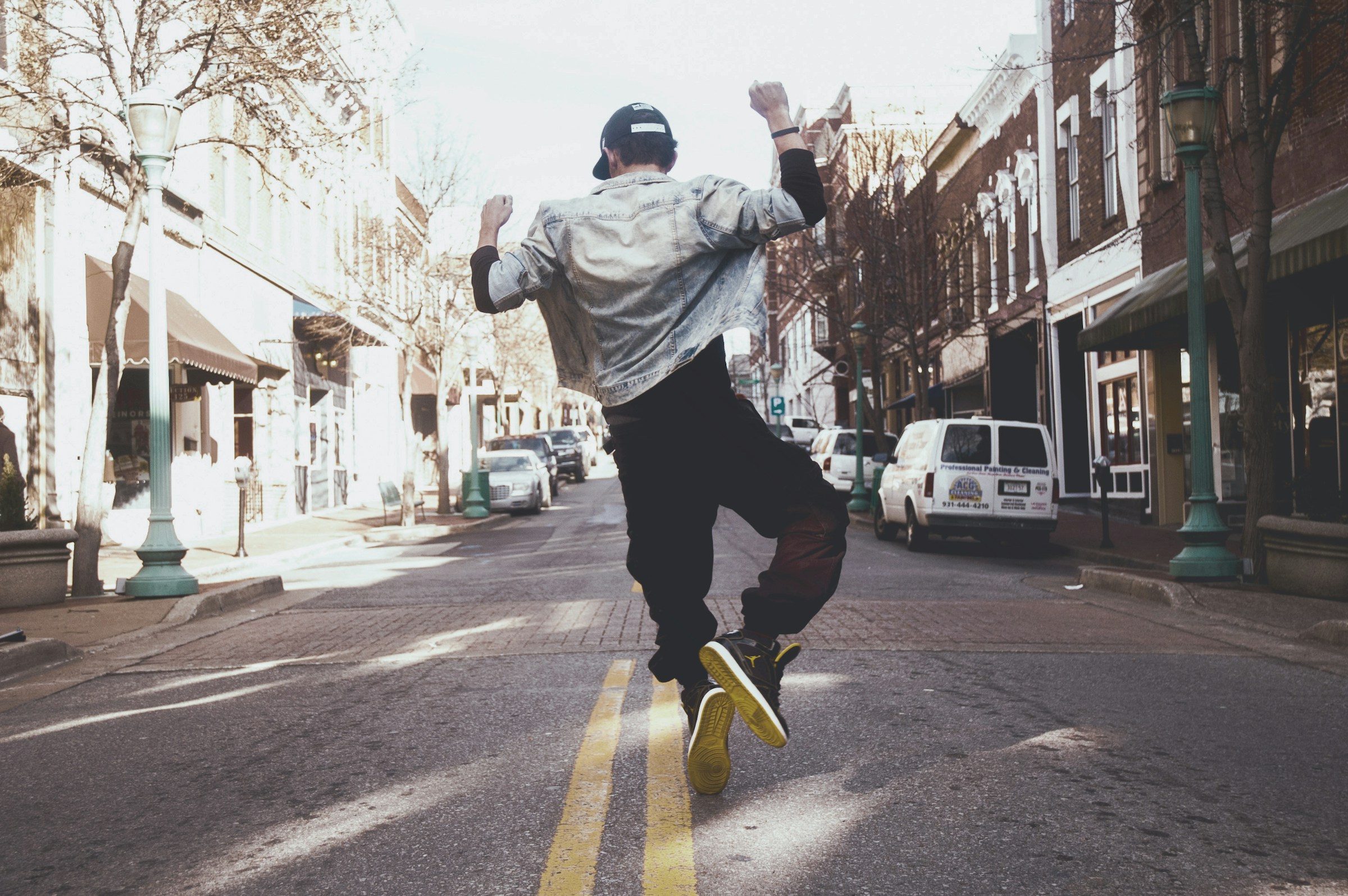 A person in a jean jacket and ball cap dances in the middle of a city street.