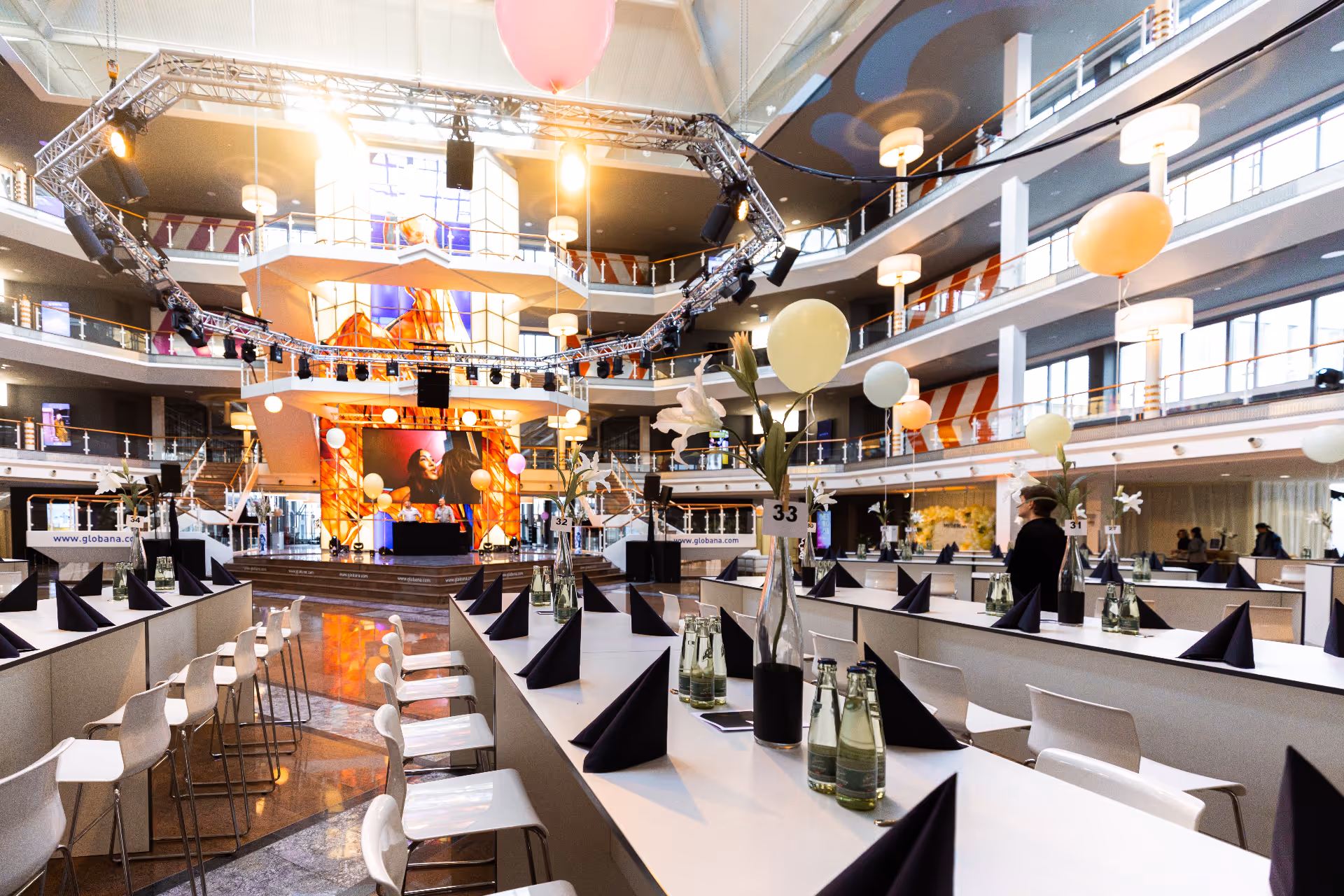 Moderner, mehrstöckiger Veranstaltungsraum mit langen weißen Tischen, gedeckt mit schwarzen Servietten, Wasserflaschen, weißen Stühlen, Blumenvasen und an Stühlen befestigten Luftballons.