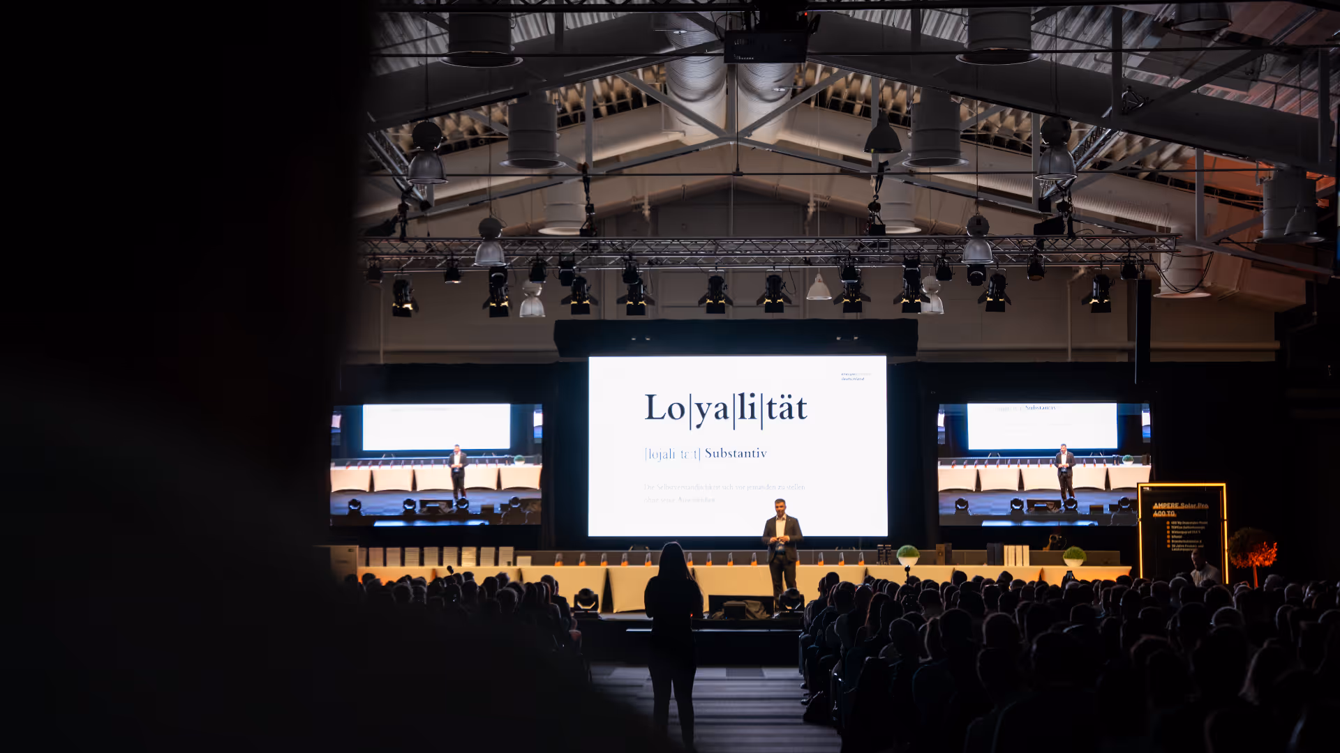 Redner auf der Bühne, der vor einem großen Publikum spricht, während auf einer zentralen Leinwand und zwei seitlichen Leinwänden in einem Konferenzsaal das Wort „Loyalität” angezeigt wird.