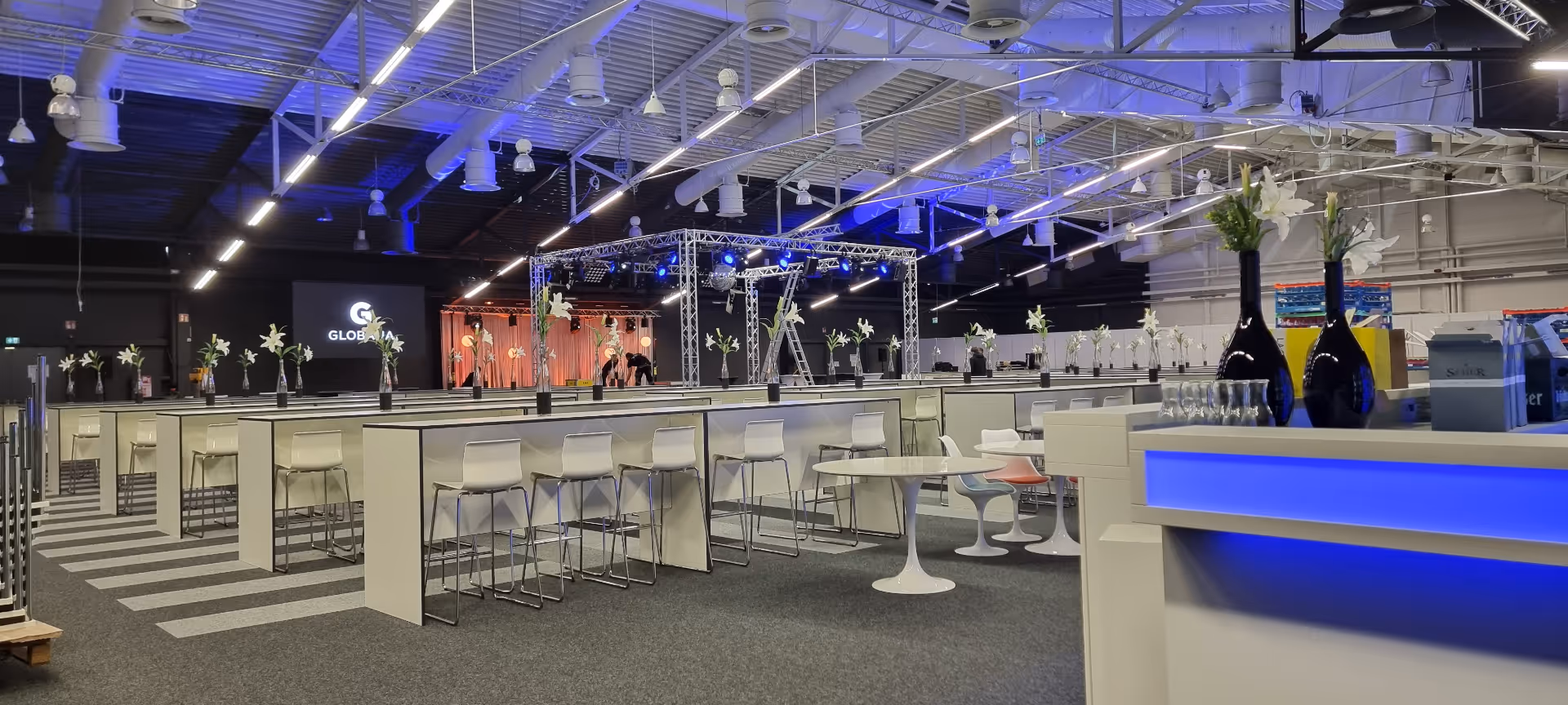 Moderner Veranstaltungsraum mit Reihen von hohen Tischen und Hockern, dekoriert mit weißen Blumenarrangements unter violetter Beleuchtung.