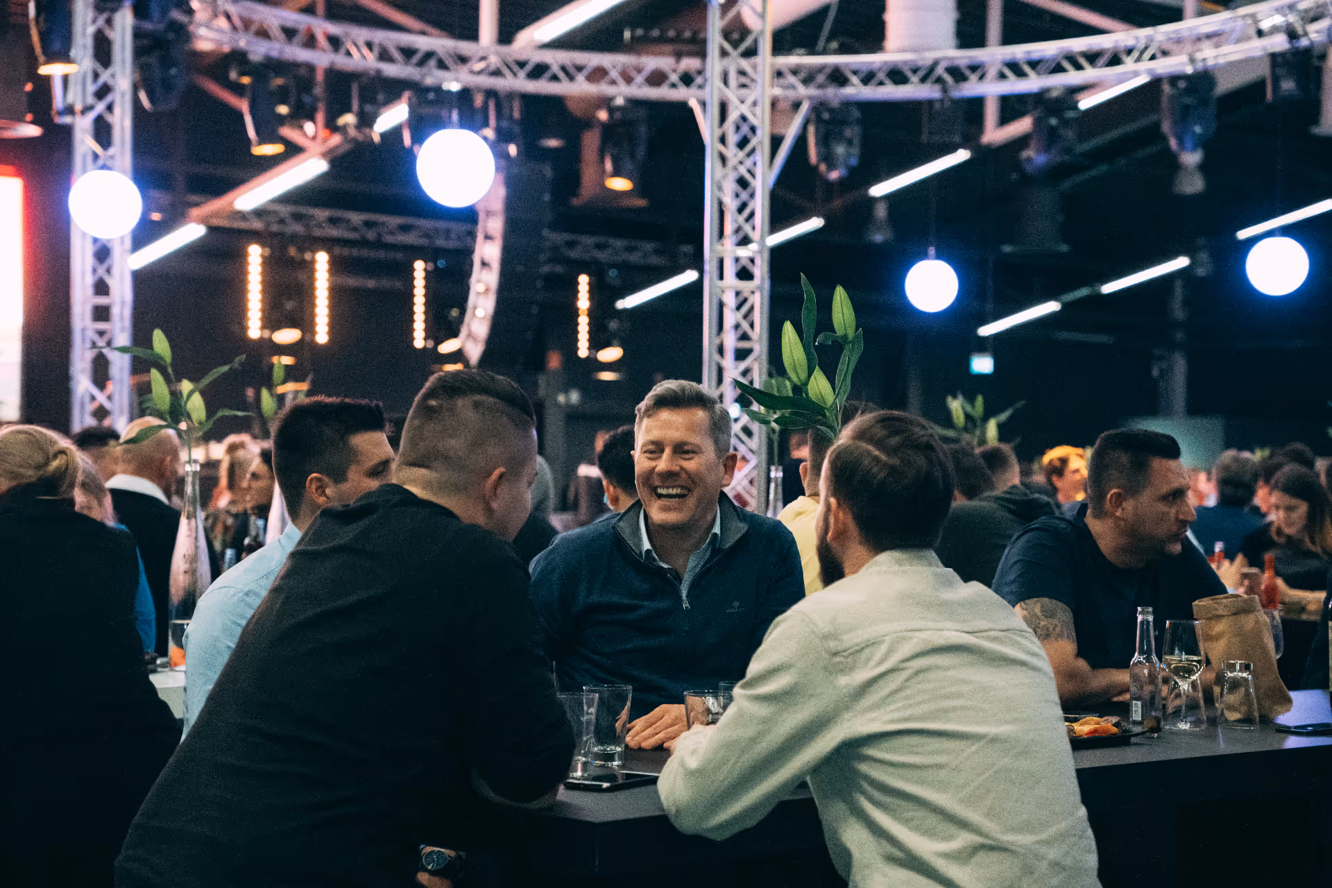 Eine Gruppe von Männern sitzt lachend um einen Tisch in einem lebhaften Veranstaltungsraum mit dekorativen Lichtern und Pflanzen.