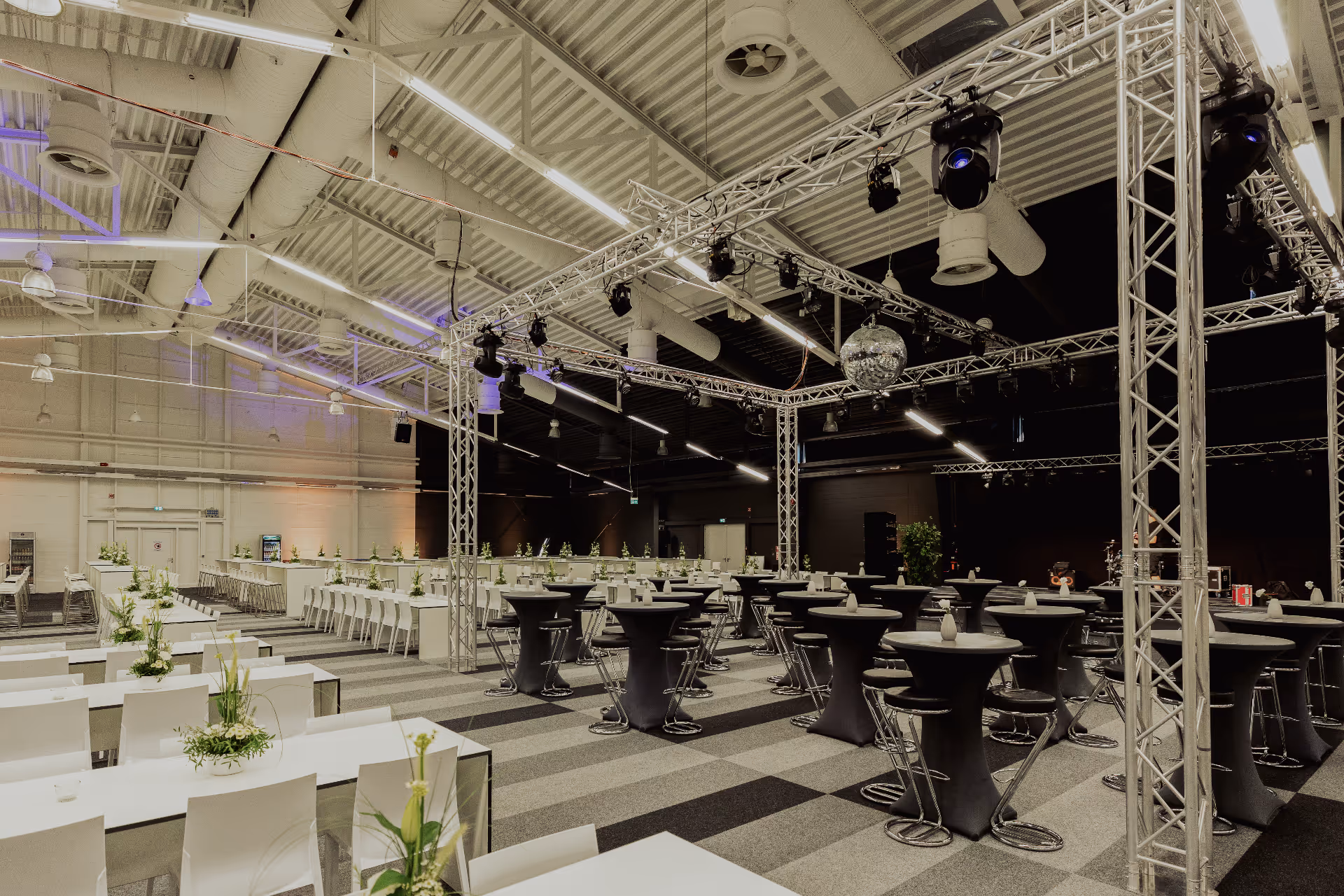 Großer Veranstaltungsraum mit weißen Stühlen und Tischen, die links mit Blumengestecken geschmückt sind, und hohen schwarzen Cocktailtischen mit Hockern rechts unter Metalltraversen und einer Discokugel an der Decke.