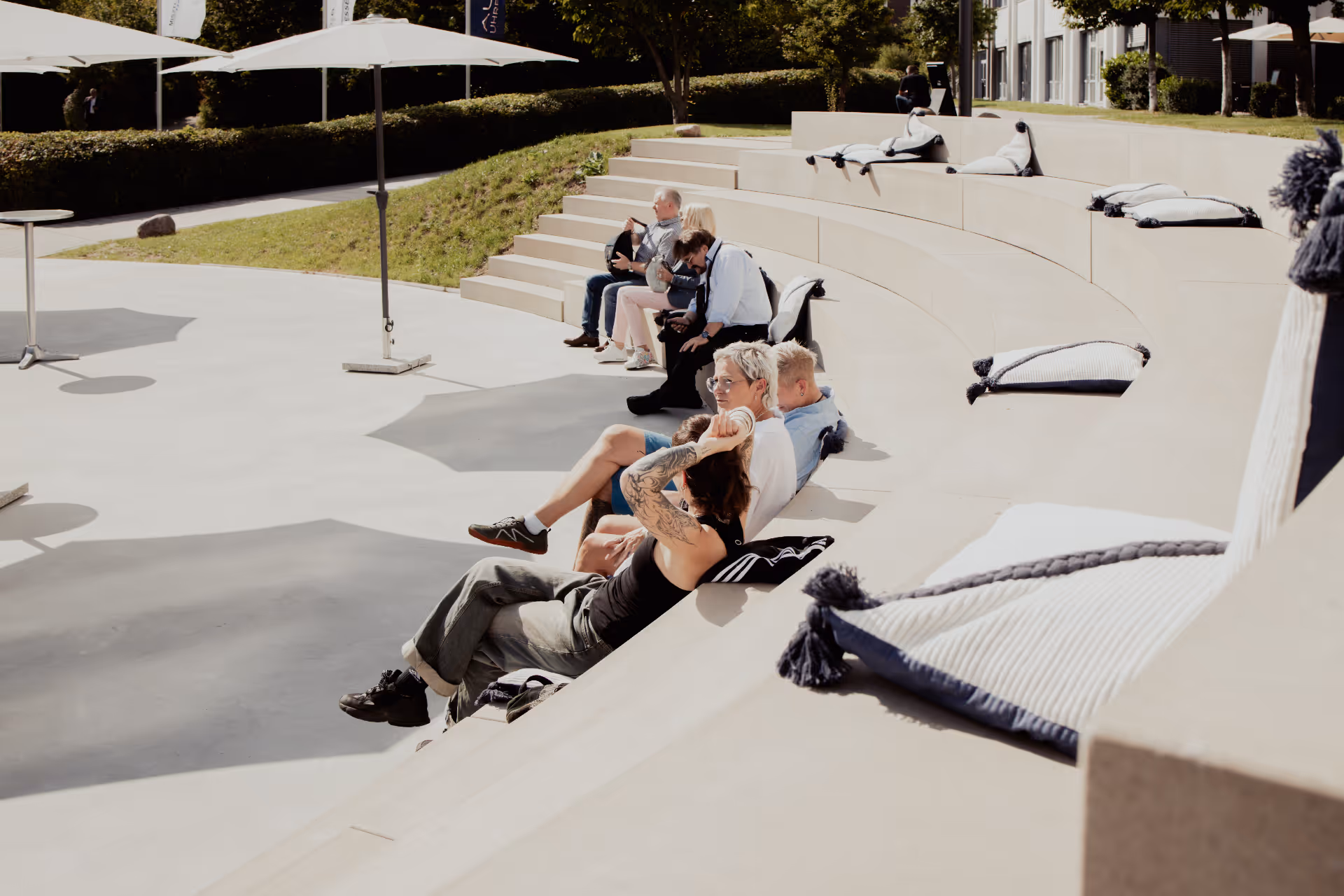 Menschen sitzen und entspannen sich an einem sonnigen Tag auf Betonstufen mit Kissen im Freien.