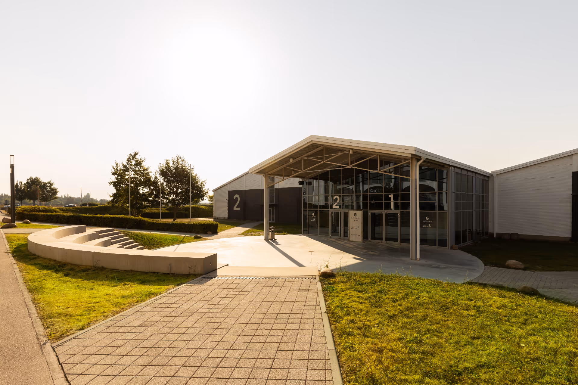 Modernes Gebäude mit großem Glaseingang, gekennzeichnet mit den Zahlen 1 und 2, umgeben von grünem Rasen und gepflastertem Gehweg unter klarem Himmel.
