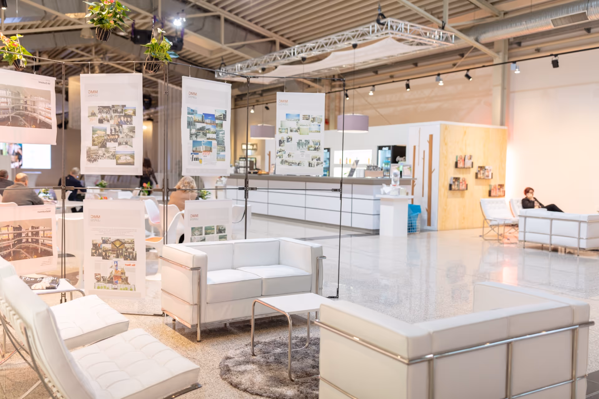 Moderner Loungebereich mit weißen Ledersofas und -sesseln, Informationspostern auf schwarzen Rahmen und einem hellen, offenen Raum mit einer Rezeption im Hintergrund.