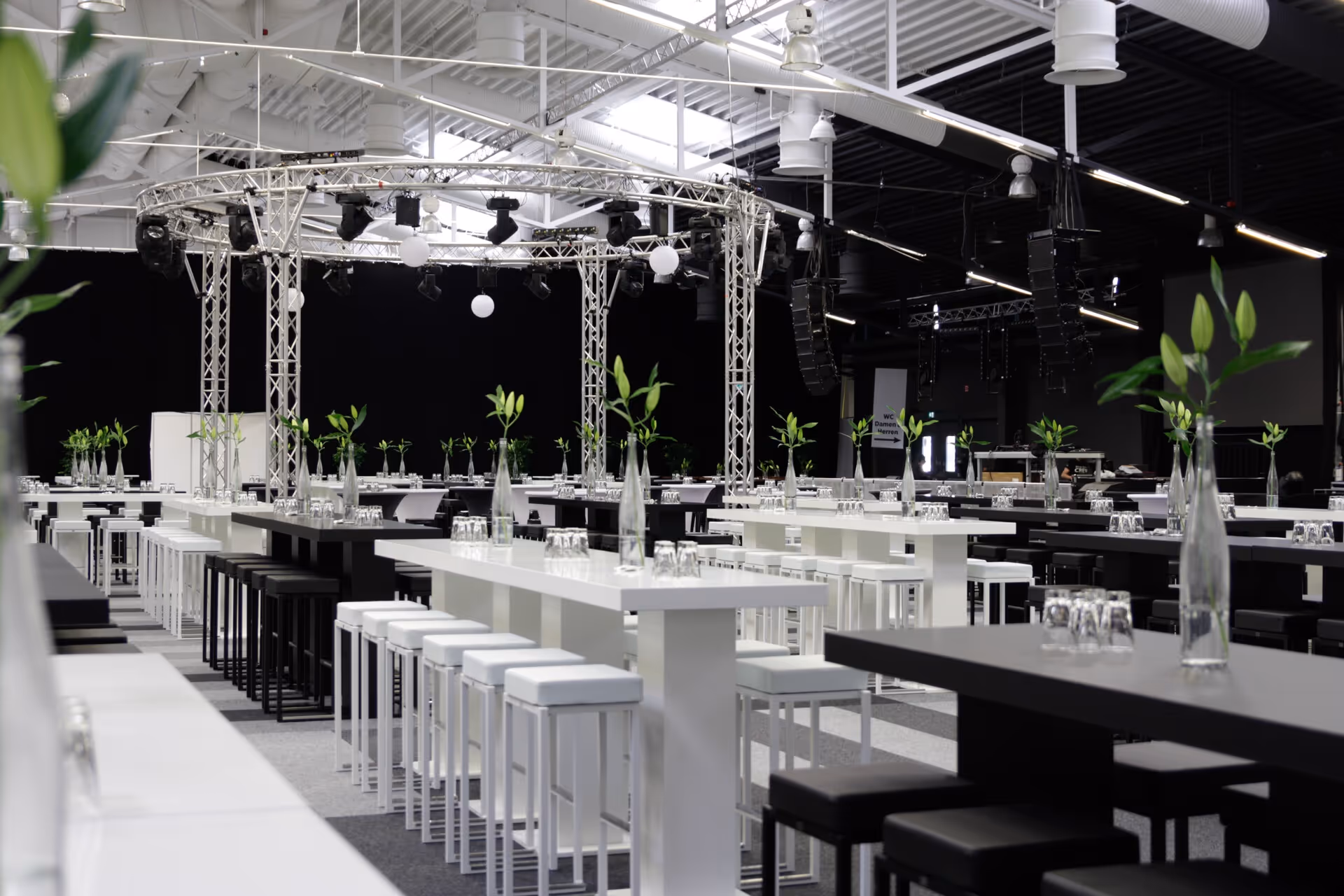 Moderner Veranstaltungsraum mit weißen und schwarzen Stehtischen und Hockern, dekoriert mit Glasflaschen mit grünen Blattstielen, unter industrieller Beleuchtung und Rigging.