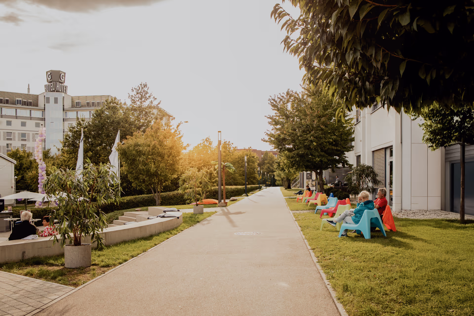 Menschen sitzen auf bunten Stühlen entlang eines gepflasterten Weges in einer grünen Außenanlage in der Nähe eines Hotelgebäudes unter einem sonnigen Himmel.