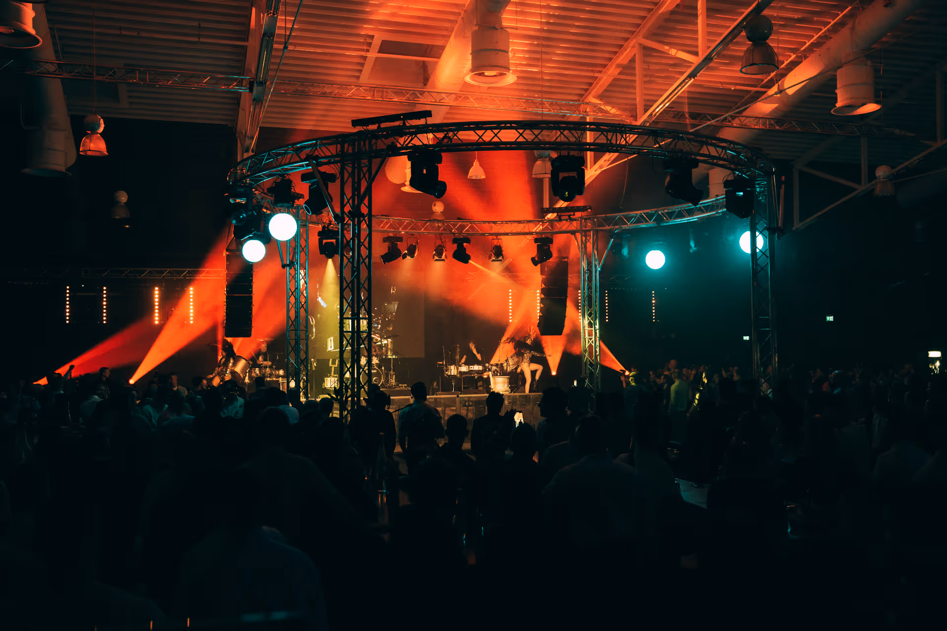 Konzertbühne mit kreisförmiger Traversenbeleuchtung und roten und blauen Scheinwerfern, die die Künstler beleuchten, mit einer dunklen Menschenmenge im Vordergrund.