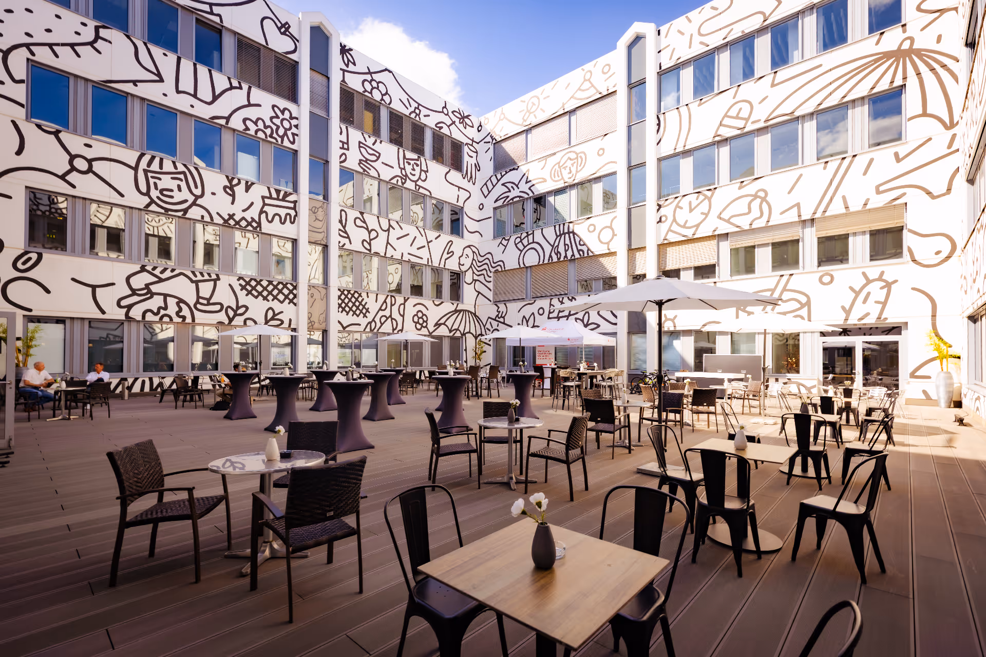 Außenhof mit Tischen, Stühlen, Sonnenschirmen und hohen Cocktailtischen vor einer Gebäudefassade, die mit großen schwarz-weißen Wandmalereien verziert ist.
