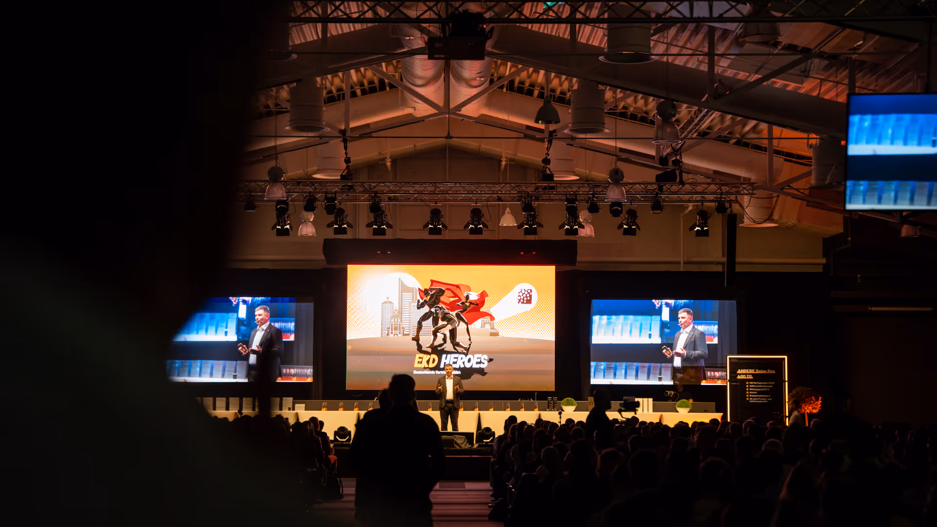 Moderator auf der Bühne spricht zu einem Publikum, während große Bildschirme ihn zeigen und ein zentraler Bildschirm die Grafik „EKD Heroes“ anzeigt.