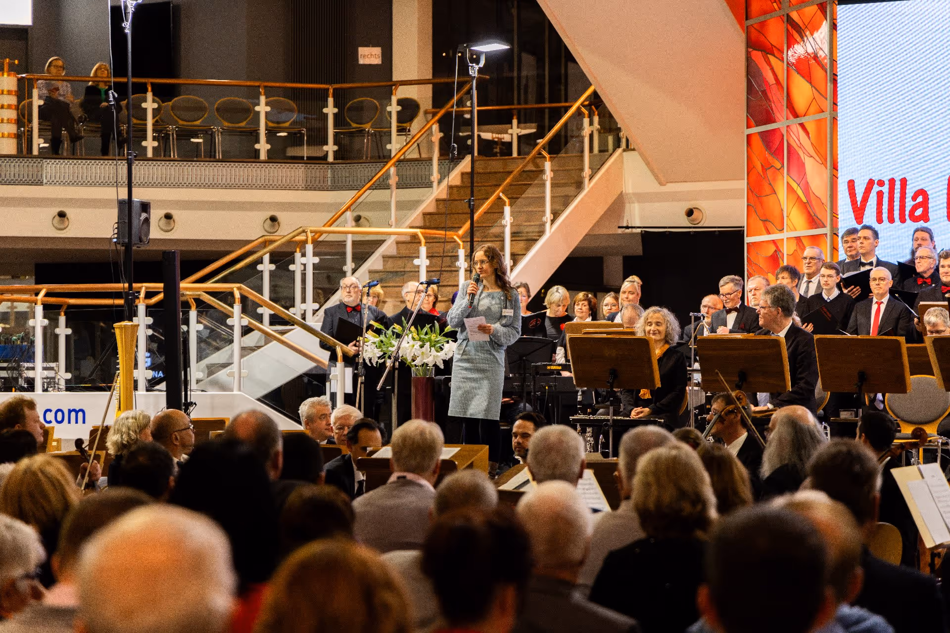 Eine Frau in einem blauen Kleid spricht vor sitzenden Musikern und einem Chor in einem Innenauditorium auf einer Bühne in ein Mikrofon.