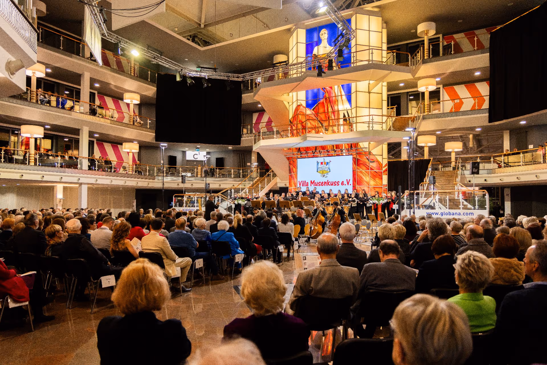 Das Publikum sitzt in einem großen, modernen Auditorium und verfolgt eine Live-Aufführung eines Orchesters und Chors auf der Bühne vor einer farbenfrohen Kulisse und einer Leinwand mit der Aufschrift „Villa Musenkuss e.V.“.