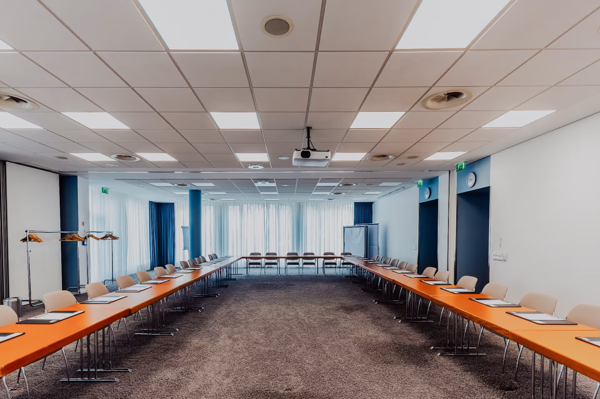 Großer U-förmiger Konferenzraum mit langen Holztischen, beigen Stühlen und einem an der Decke montierten Projektor.