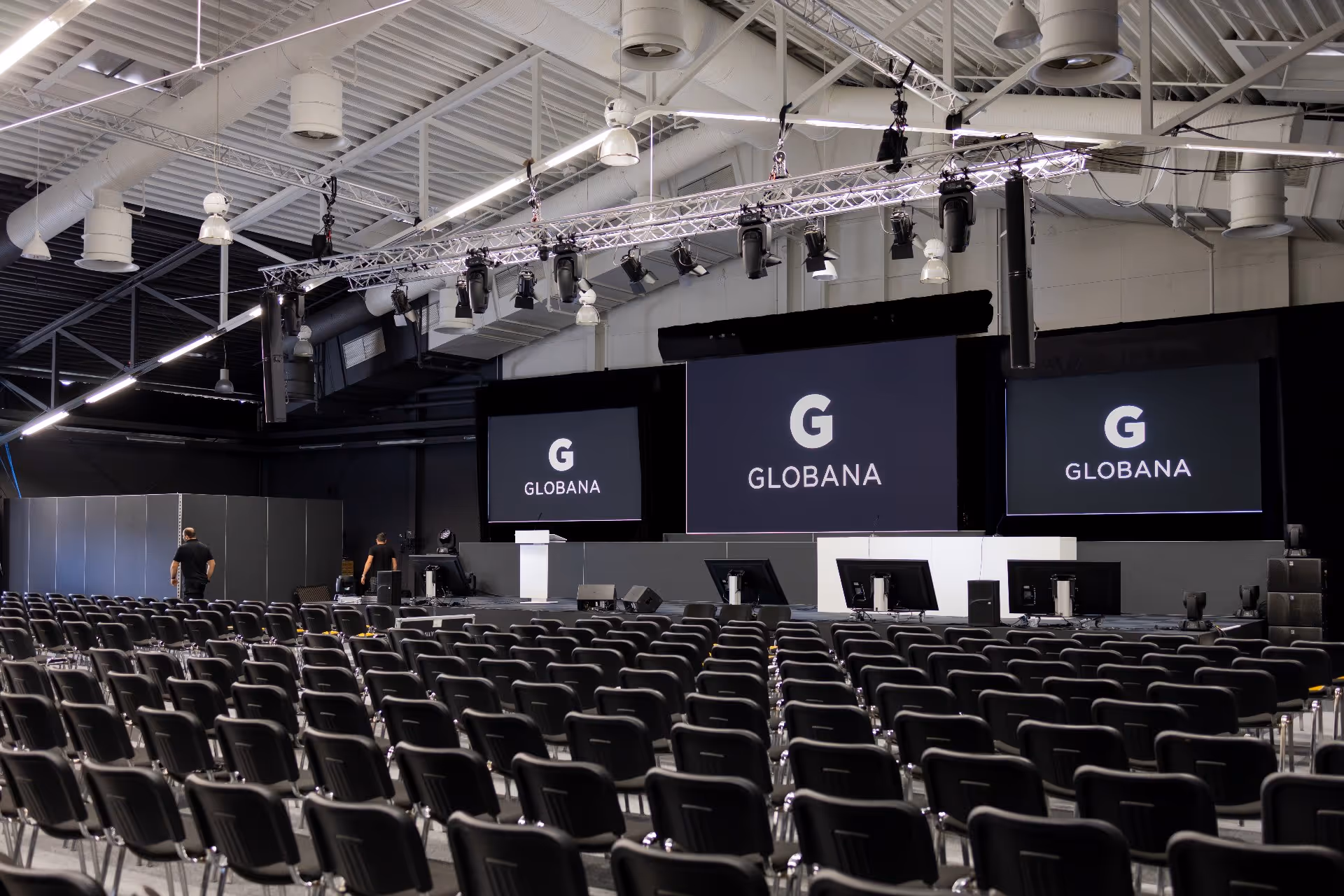 Leerer Konferenzraum mit Reihen schwarzer Stühle, die auf eine Bühne mit drei großen Bildschirmen ausgerichtet sind, auf denen das Wort „GLOBANA” zu sehen ist.
