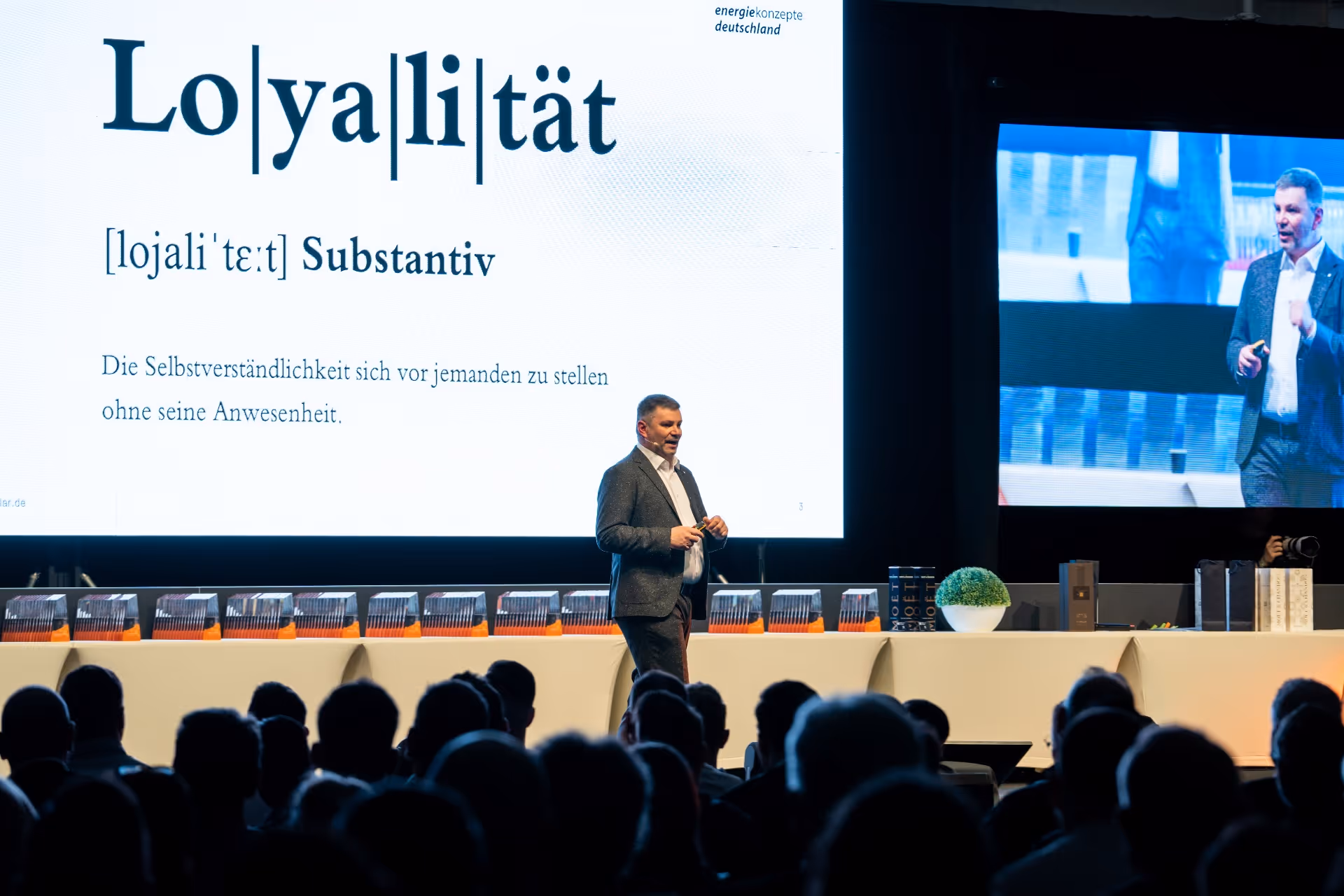 Mann hält eine Präsentation auf der Bühne, während auf einer großen Leinwand das deutsche Wort „Loyalität” und dessen Definition zu sehen sind und das Publikum im Vordergrund zusieht.