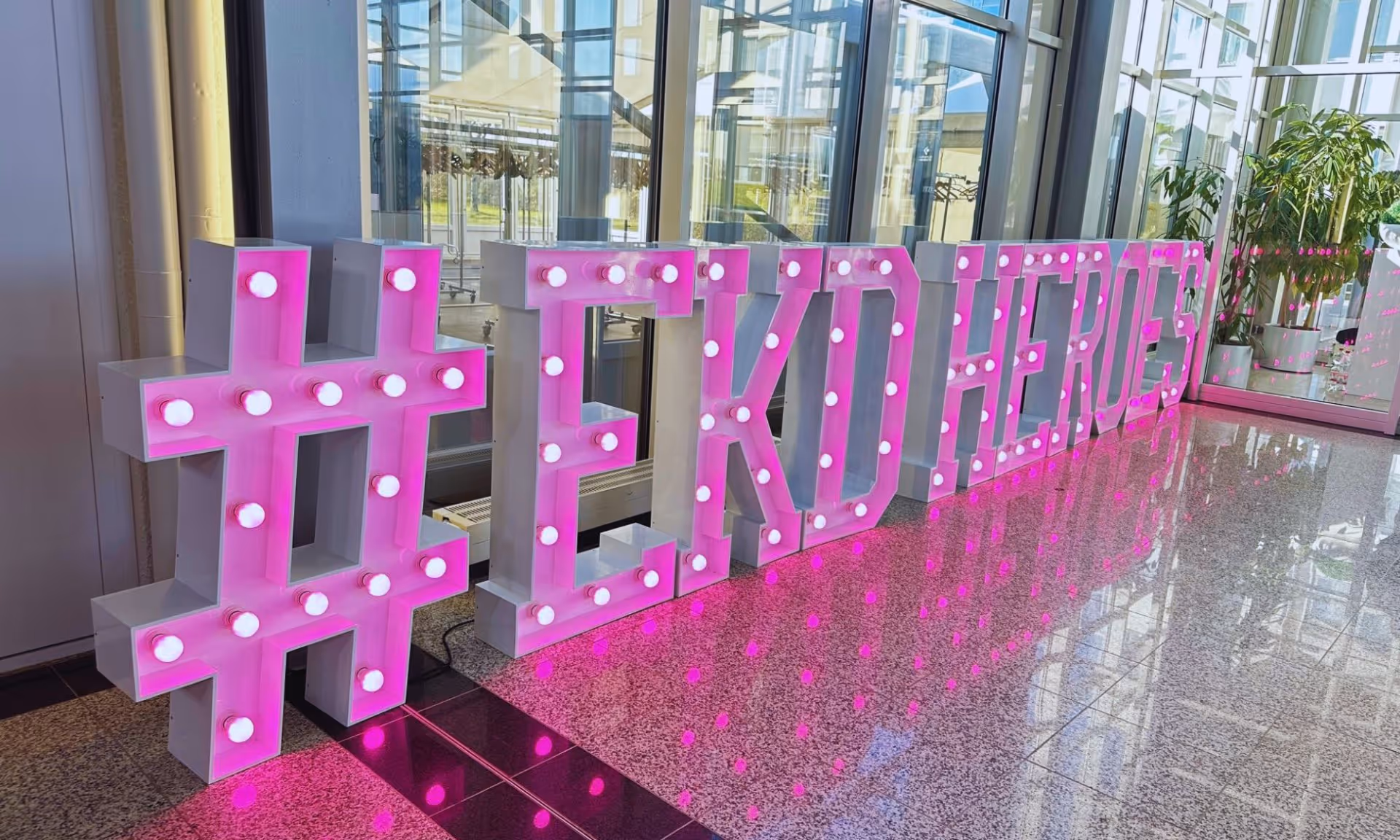 Große beleuchtete rosa Leuchtschriftbuchstaben, die den Schriftzug „#TEDxHEROES” bilden, in einem modernen Glasgebäude mit reflektierendem Bodenbelag.