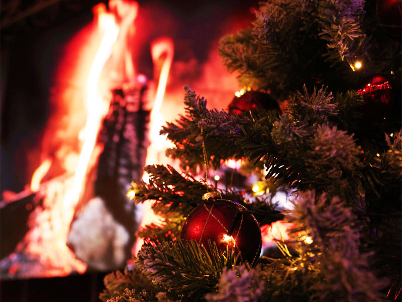 Weihnachtsbaum mit roten Kugeln und Lichterketten vor knisterndem Kaminfeuer.