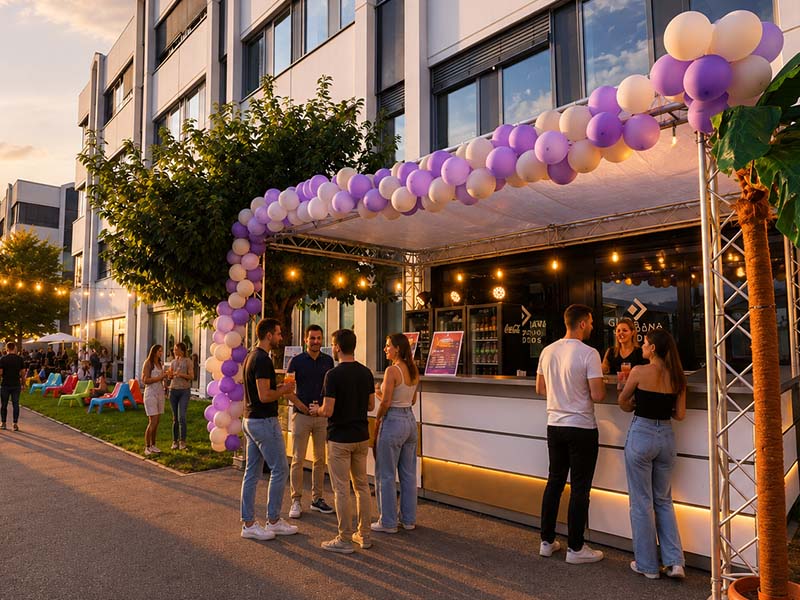 Menschen unter einem Lila-weißen Luftballonbogen vor einem Außenverkaufsstand bei Sonnenuntergang.