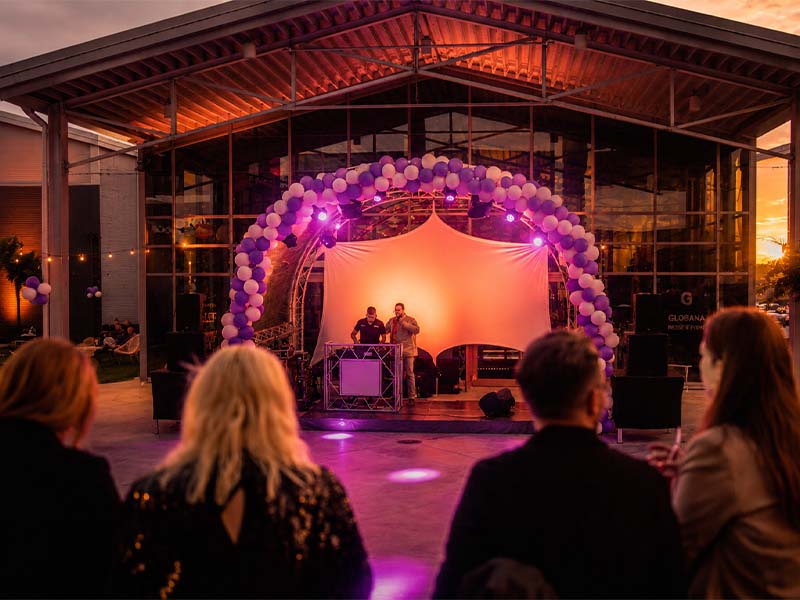 Personen sehen eine Bühnenperformance mit leisem Licht und Luftballonbogen im Freien.