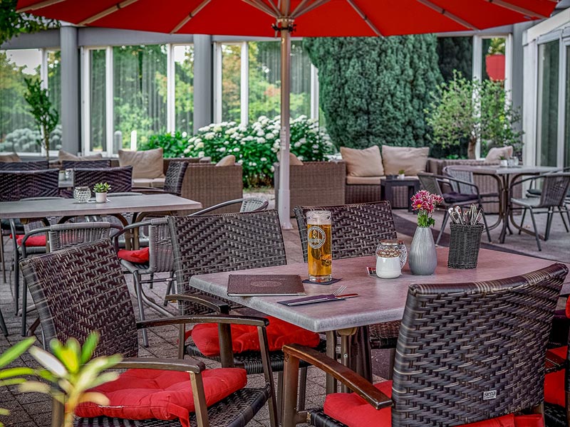 Restaurant-Außenterrasse mit Tischen, Stühlen mit roten Kissen, einem Bier und Blumenvase unter einem roten Sonnenschirm.