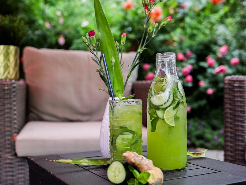 Grünes Getränk mit Gurke und Limette in Glas und Flasche auf Gartentisch neben Ingwer und Pflanze.