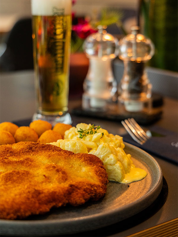 Teller mit paniertem Schnitzel, Käsespätzle und Kartoffelbällchen, im Hintergrund Glas Bier.