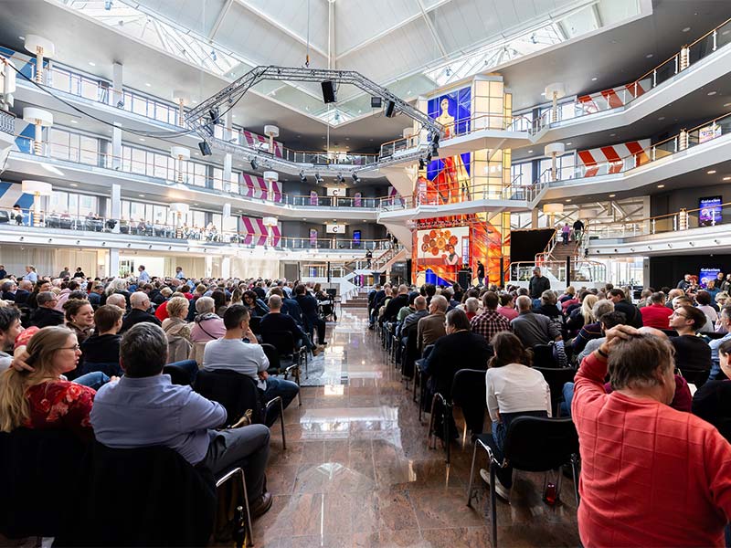 Große Menschenmenge sitzt in modernem Atrium bei einer Präsentation mit Redner auf Bühne.