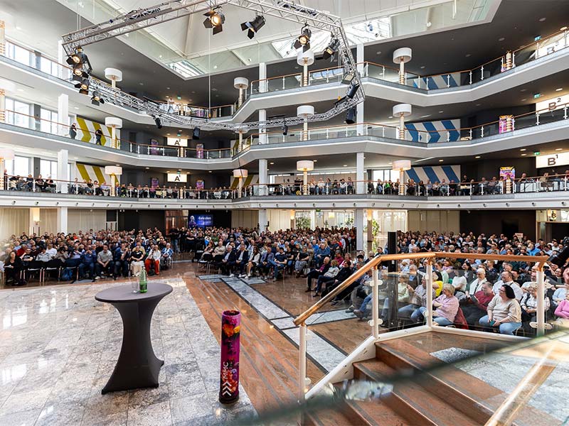Großes Auditorium mit Publikum auf Sitzplätzen und Balkonen in modernem, mehrstöckigem Veranstaltungsraum.