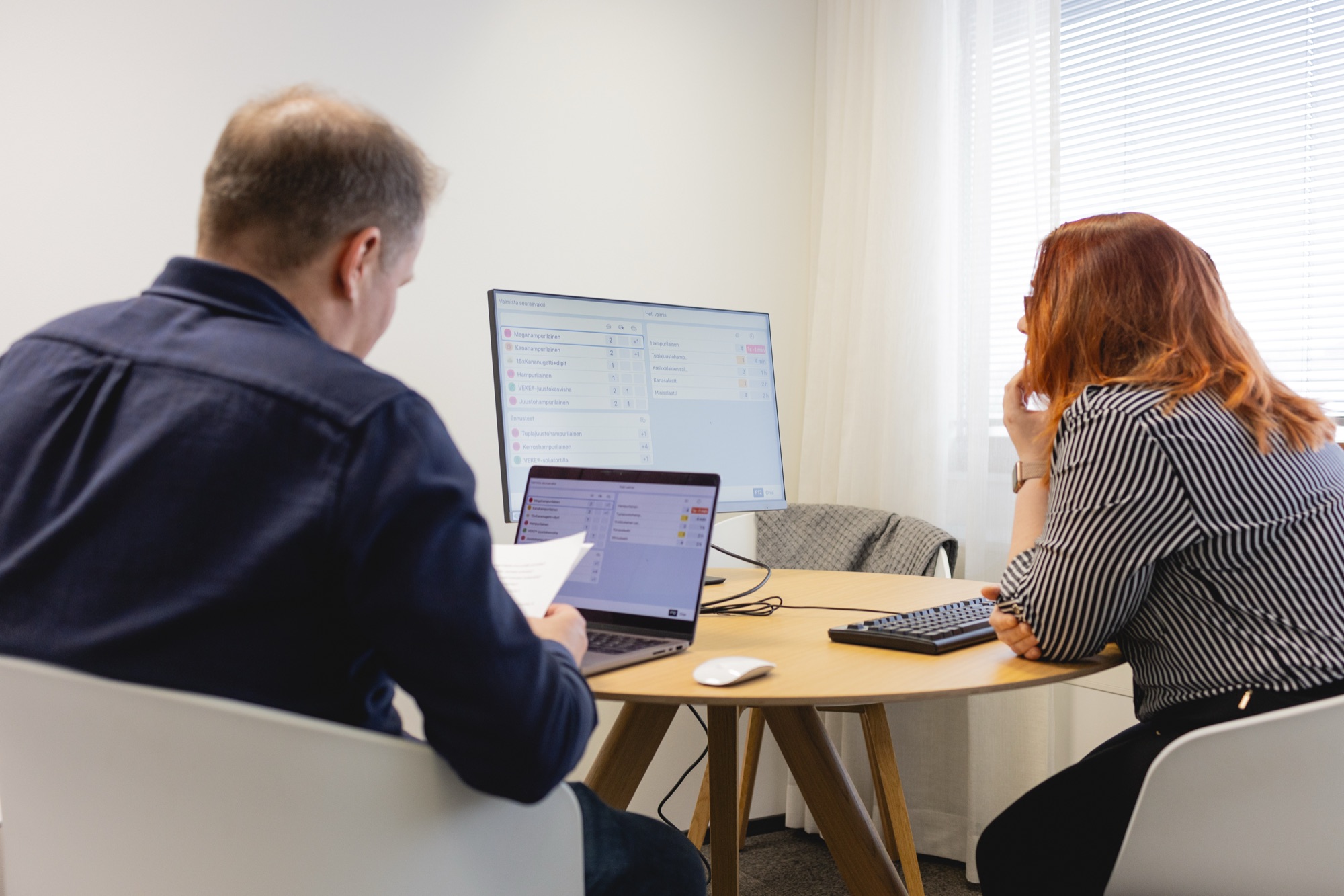 Kaksi henkilöä työskentelee kannettavan ja näytön ääressä neuvotteluhuoneessa
