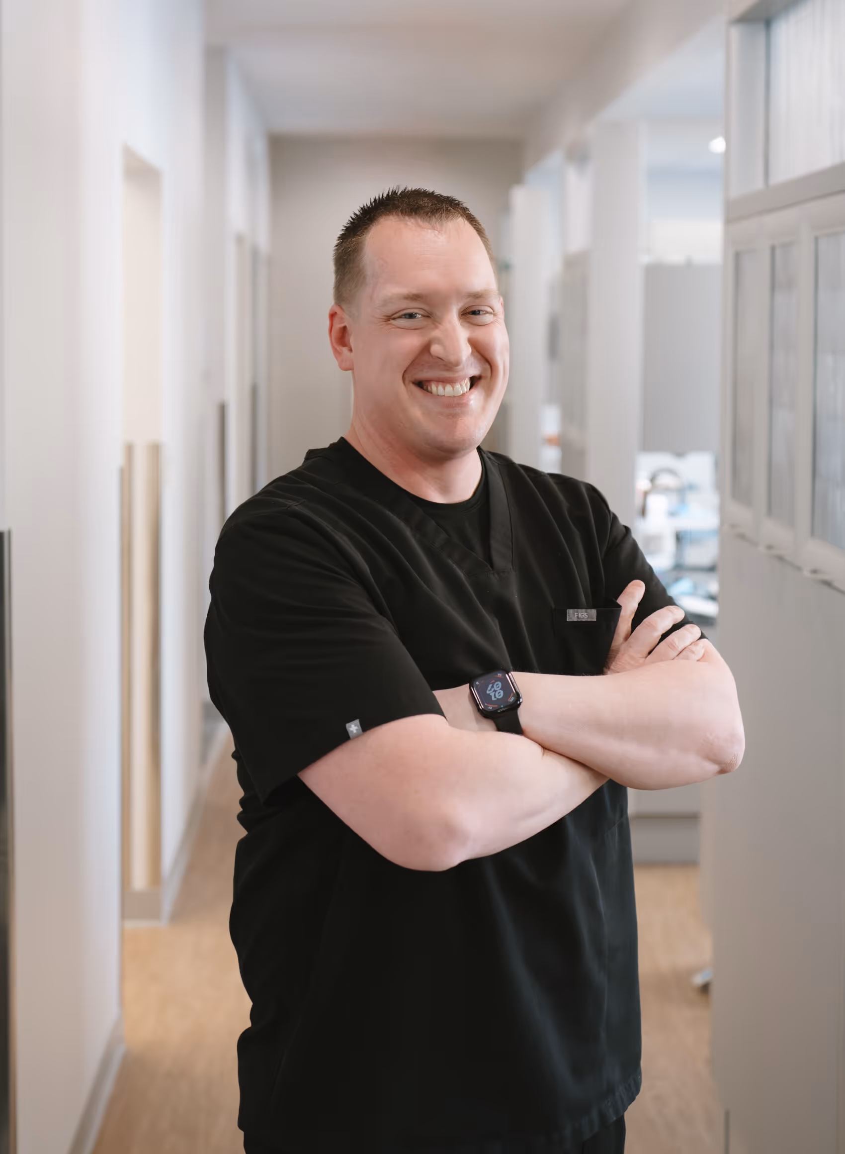 Smiling man in black medical scrubs with arms crossed standing in a bright hallway.