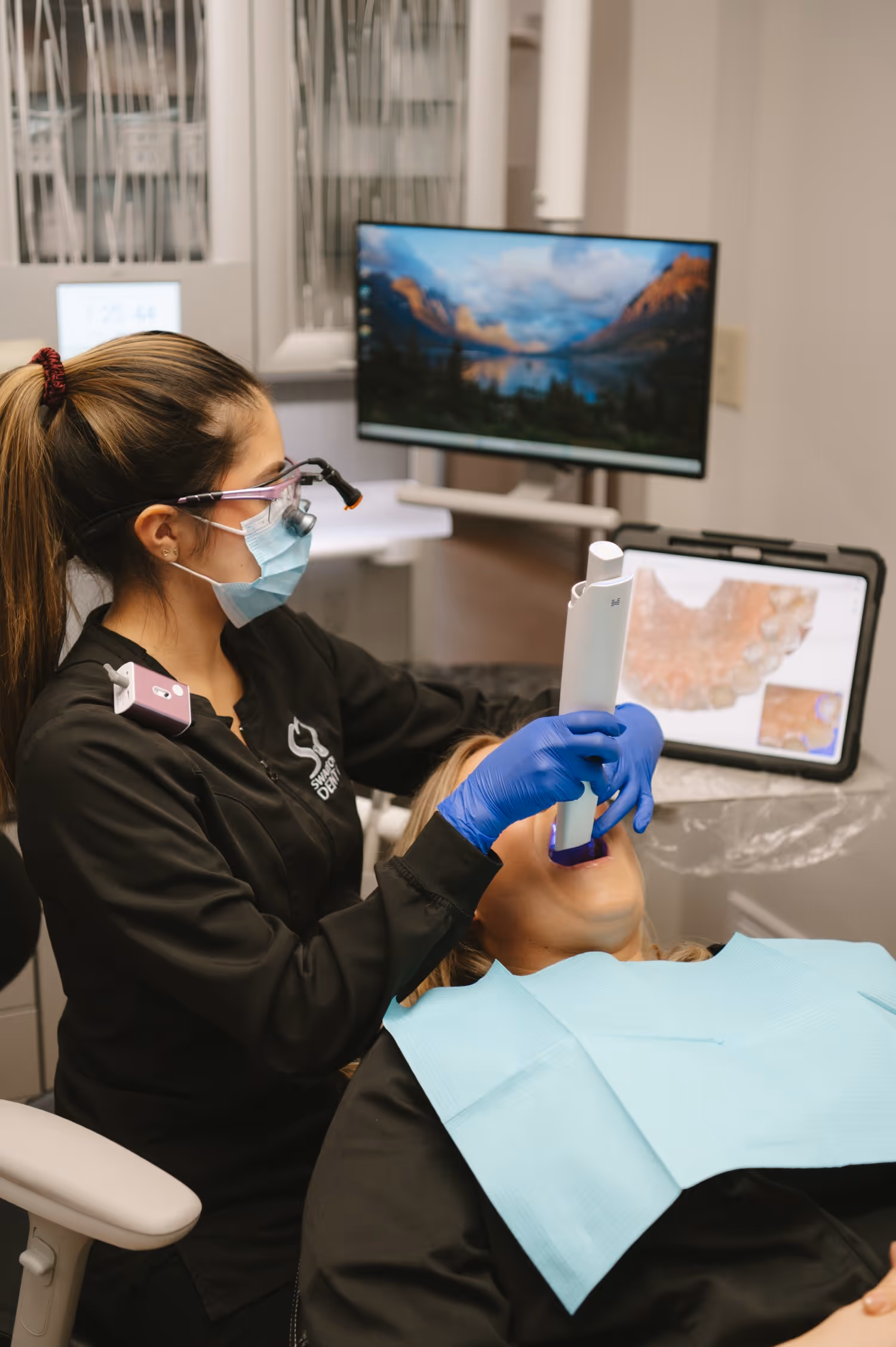 Dentist wearing a mask and magnifying glasses scanning a patient’s mouth with a handheld digital dental scanner while the scan appears on a computer screen.